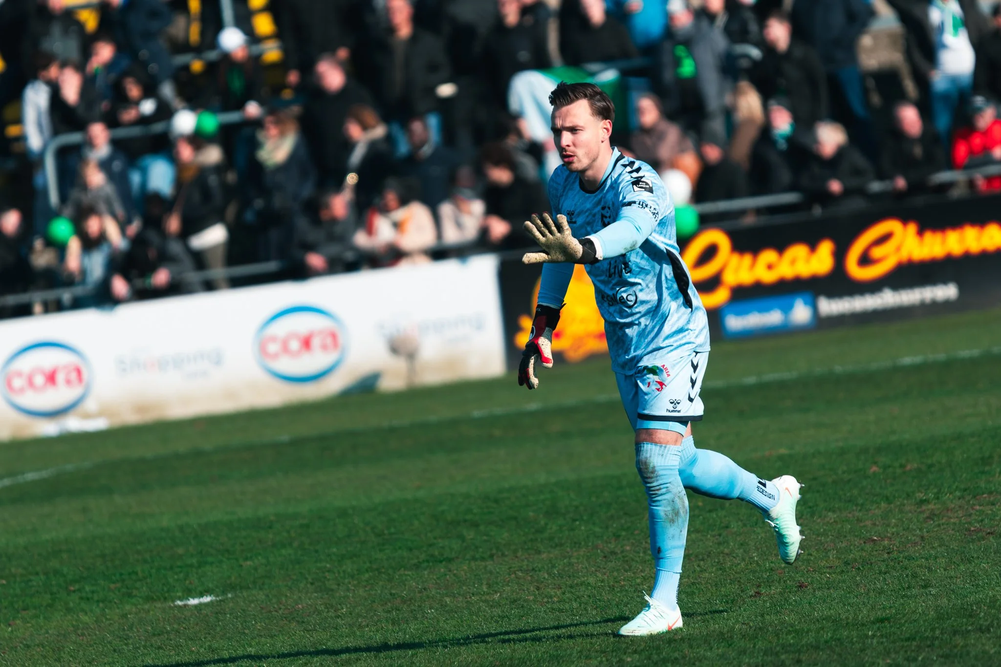 Un gardien de but en tenue bleue sur un terrain de football lors d'un match, avec des supporters en arrière-plan.