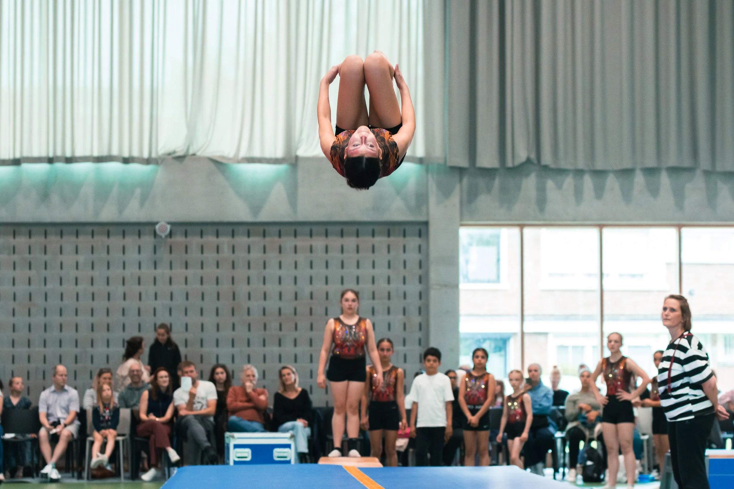 Une jeune fille effectuant une acrobatie de gymnastique dans un gymnase, avec un groupe de spectateurs et une autre gymnaste en arrière-plan.