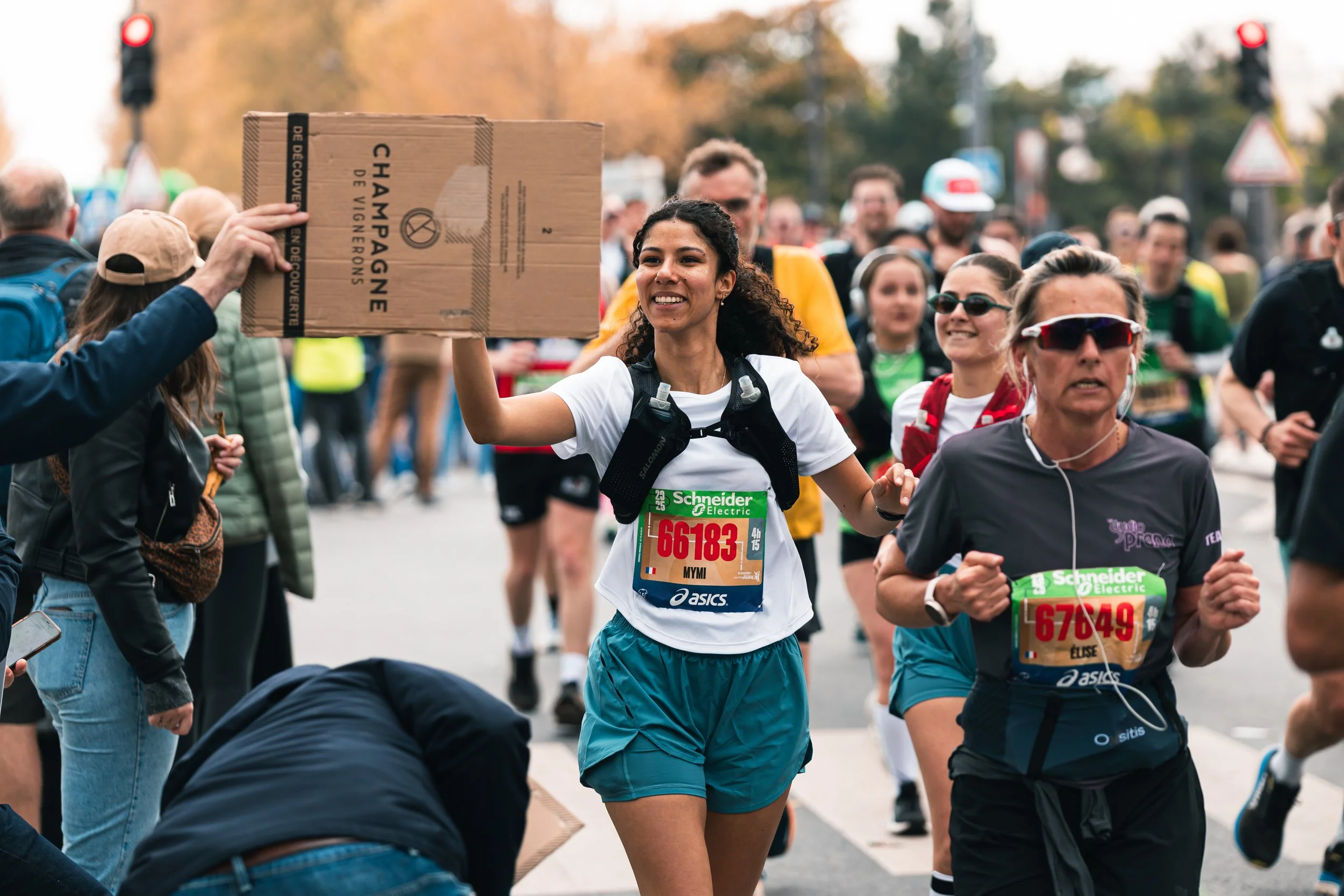 Des coureurs participent à une course de marathon, avec une femme souriante qui tend une boisson à un bénévole, dans une foule de participants.