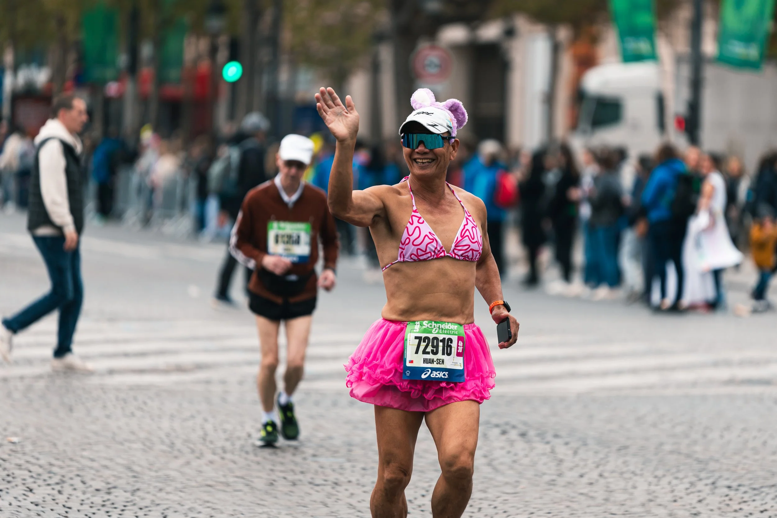 Une femme participe à une course, portant un bonnet avec des oreilles, une jupe rose à volants, et une brassière rose, souriante et saluant le public. Construits urbains en arrière-plan.