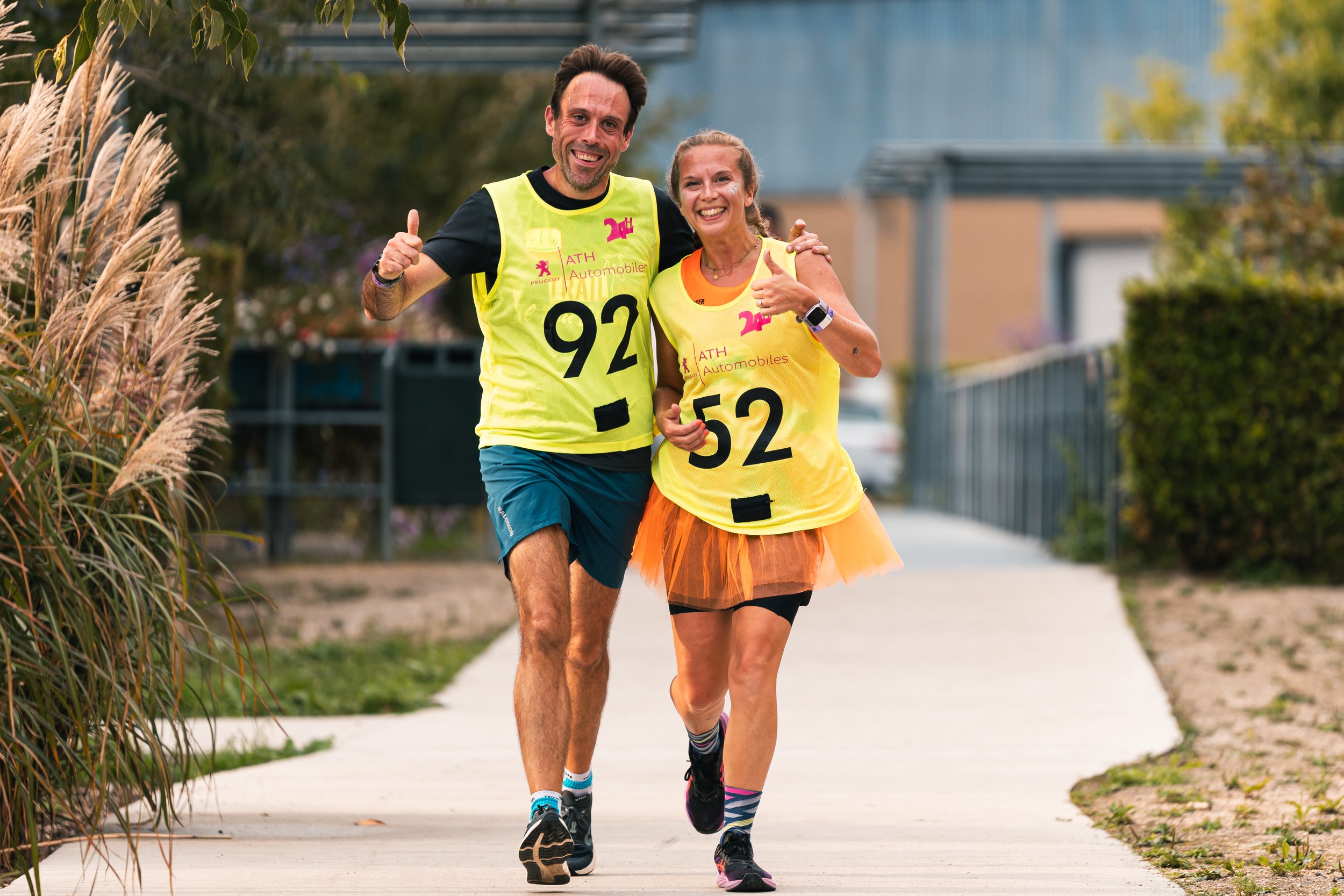 Deux coureurs en tenue de sport jaune souriants, faisant un signe de pouce en l'air, courent sur un trottoir dans un parc ou une zone résidentielle.