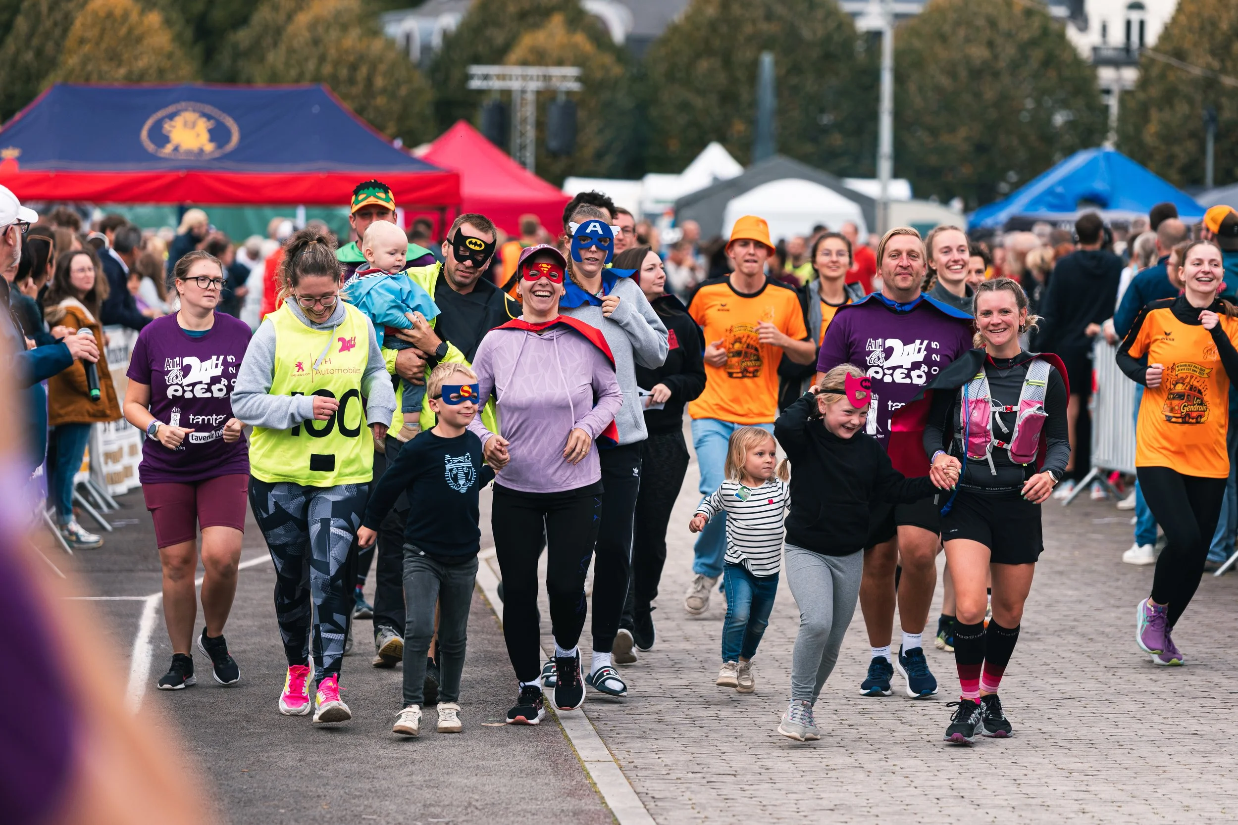 Groupe de personnes, enfants et adultes, déguisés en super-héros, participant à une marche ou course caritative dans un événement en plein air, avec des tentes colorées en arrière-plan.