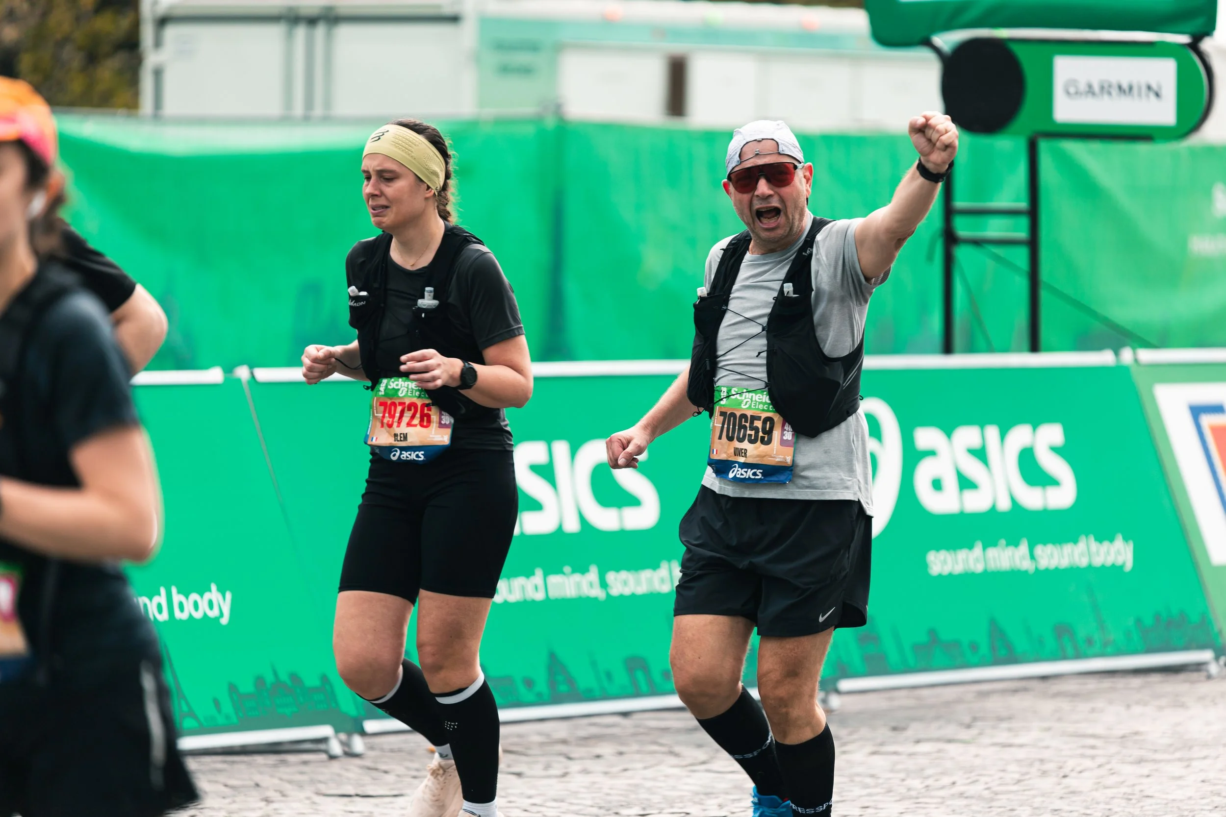 Deux coureurs participant à une course, portant des dossards et des équipements de course. L'homme porte une casquette, des lunettes de soleil, un t-shirt gris, un sac à dos, et semble encourager avec un geste de la main. La femme porte un bandeau, u
