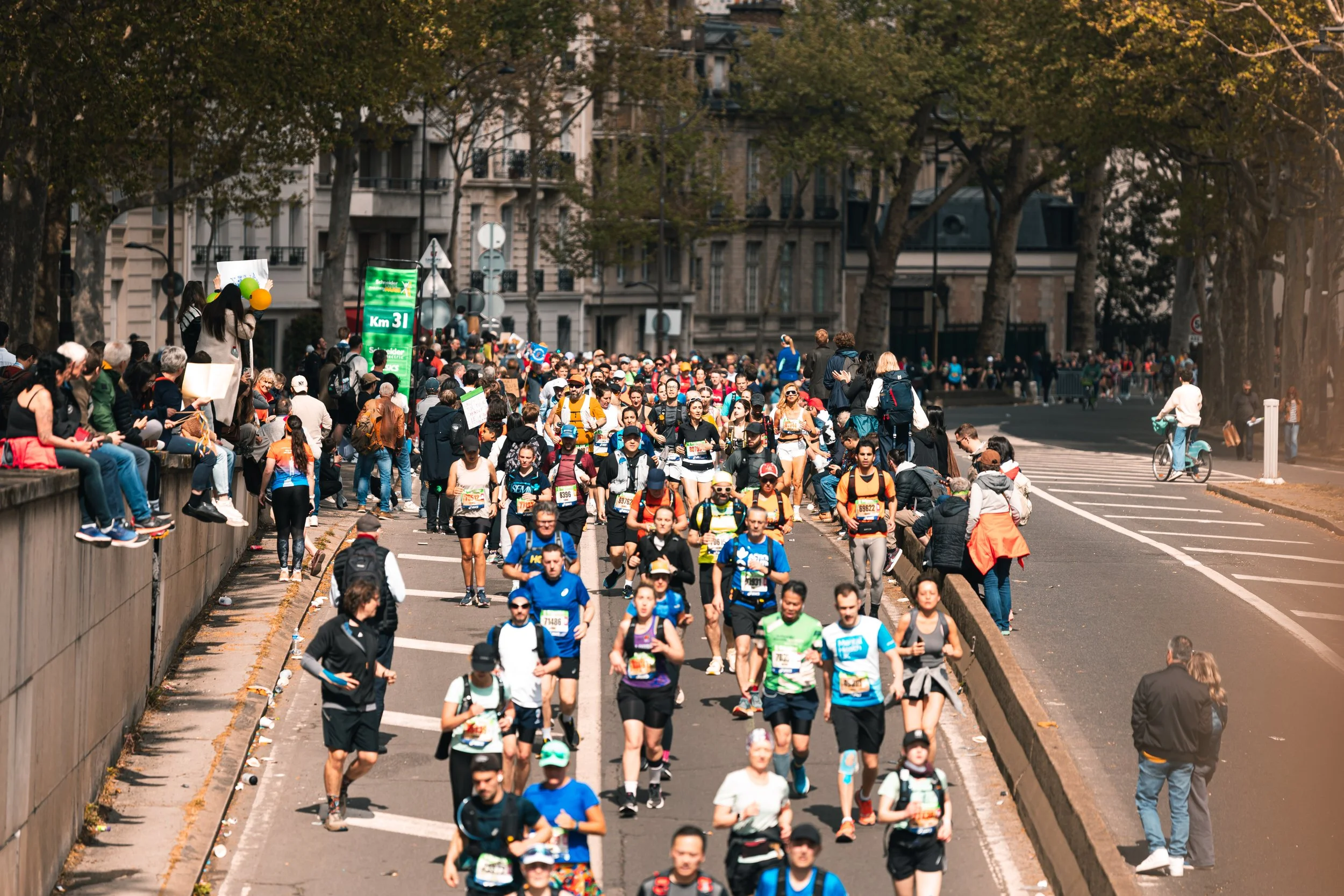 Marathon avec de nombreux coureurs et spectateurs dans une ville, arbres en arrière-plan, le matin.