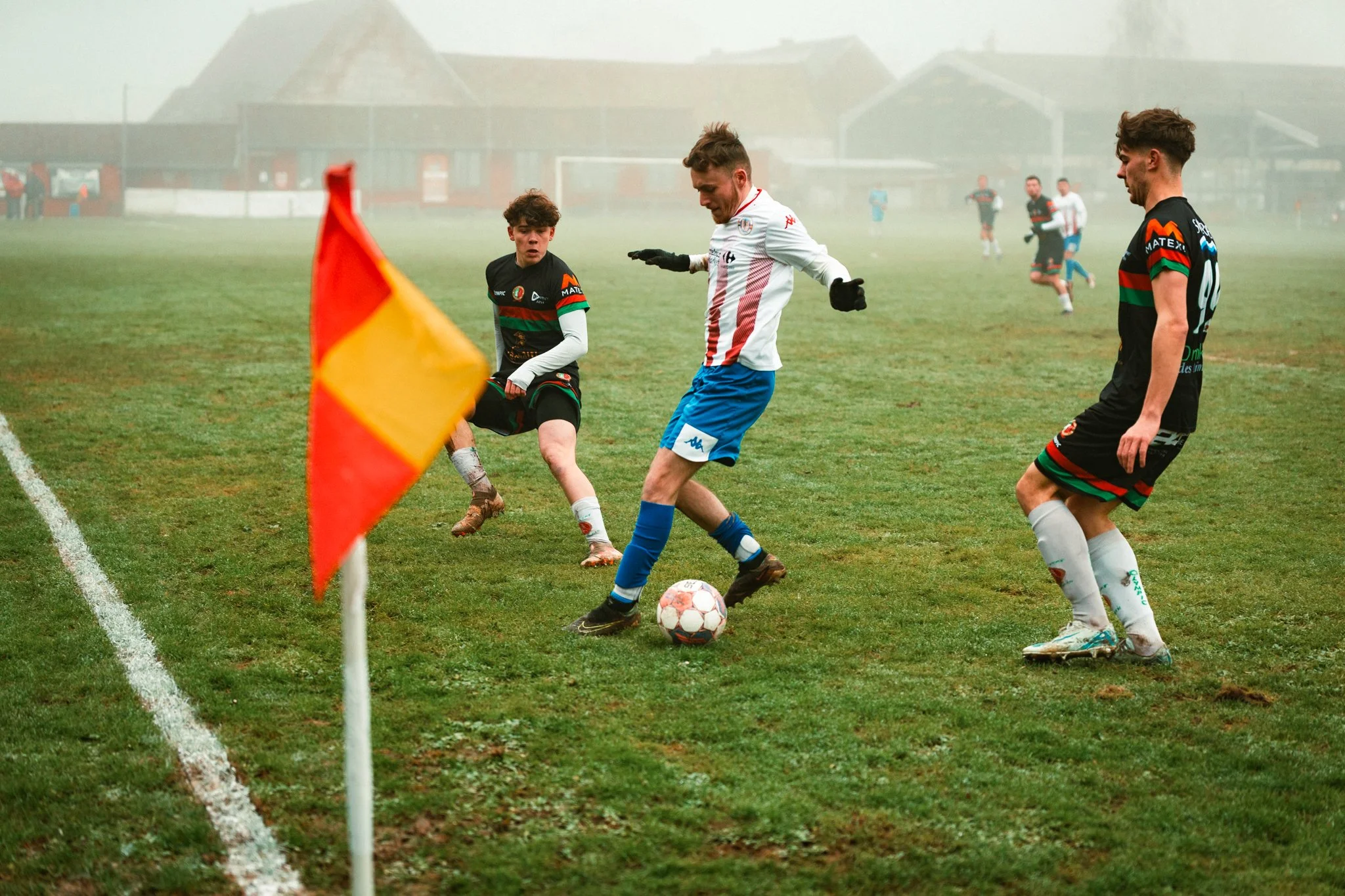 Jeunes joueurs de football en compétition sur un terrain herbeux sous un ciel brumeux, avec une balise en premier plan.