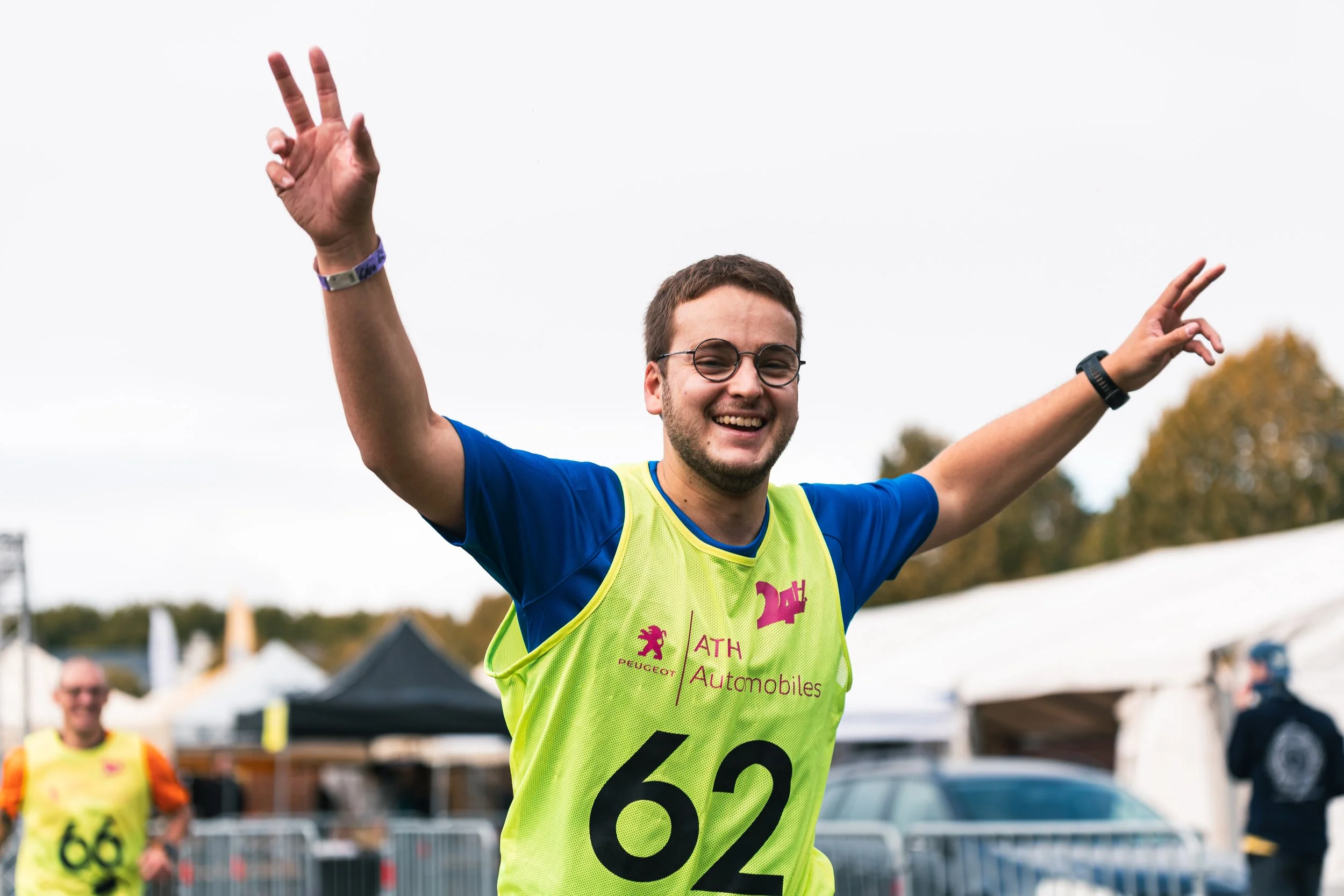 Un homme souriant avec des lunettes et un maillot jaune portant le numéro 62, levant les bras en signe de victoire lors d'une course en plein air.