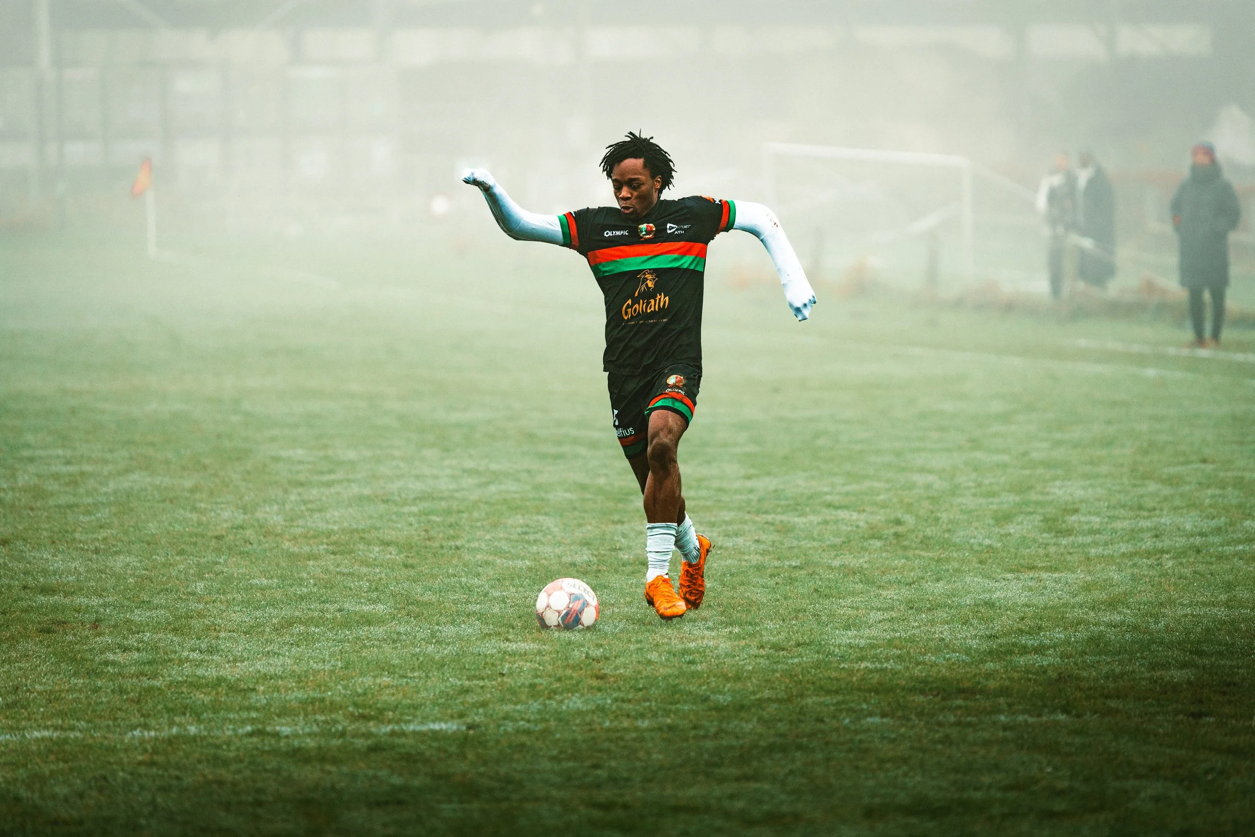 Un joueur de football en tenue noire avec des accents rouges et verts, portant des vêtements de sport orange, joue avec un ballon sur un terrain de football en plein air par temps brumeux.