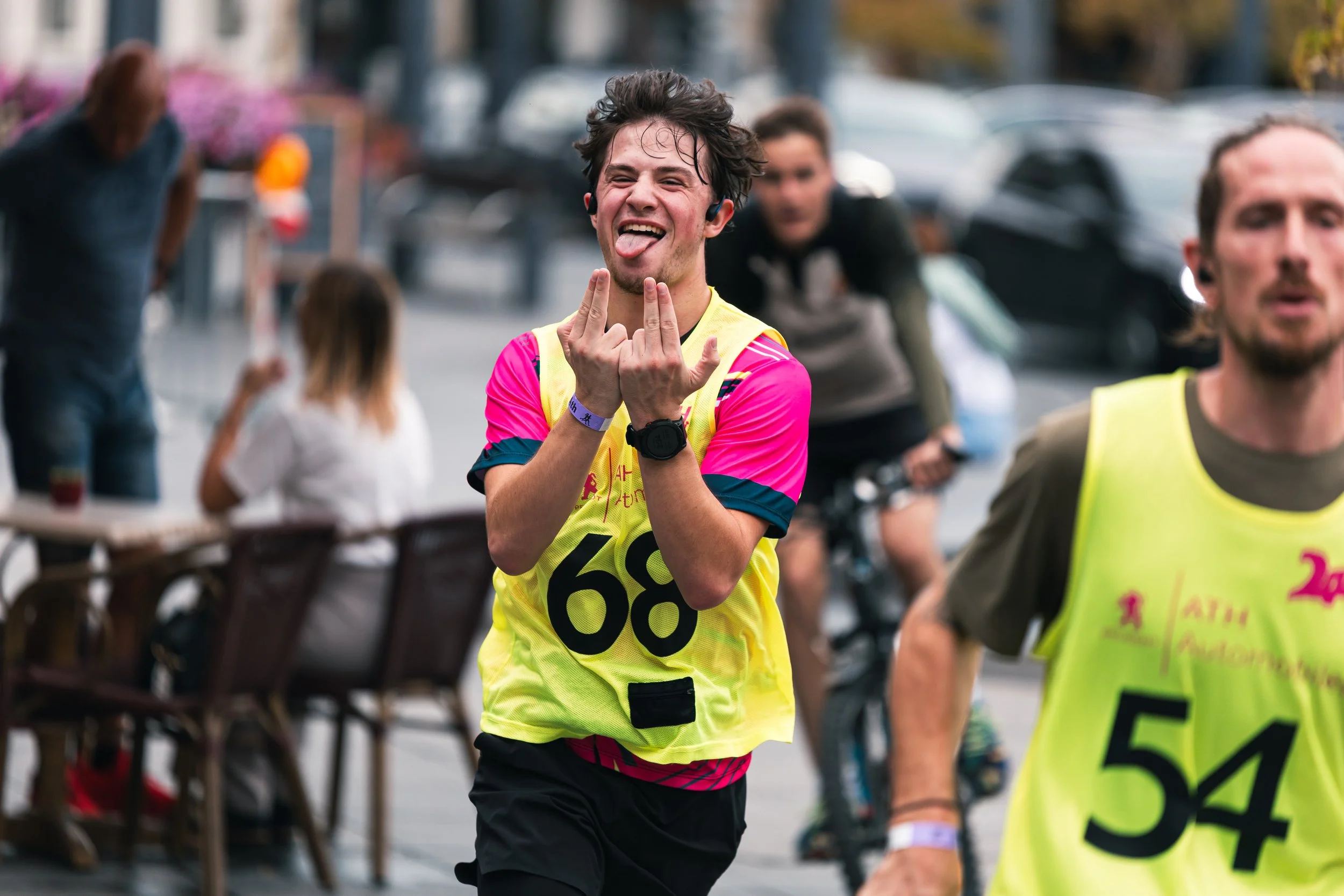 Un coureur en maillot jaune et rose faisant un geste avec ses mains lors d'une course, d'autres participants et spectateurs en arrière-plan.
