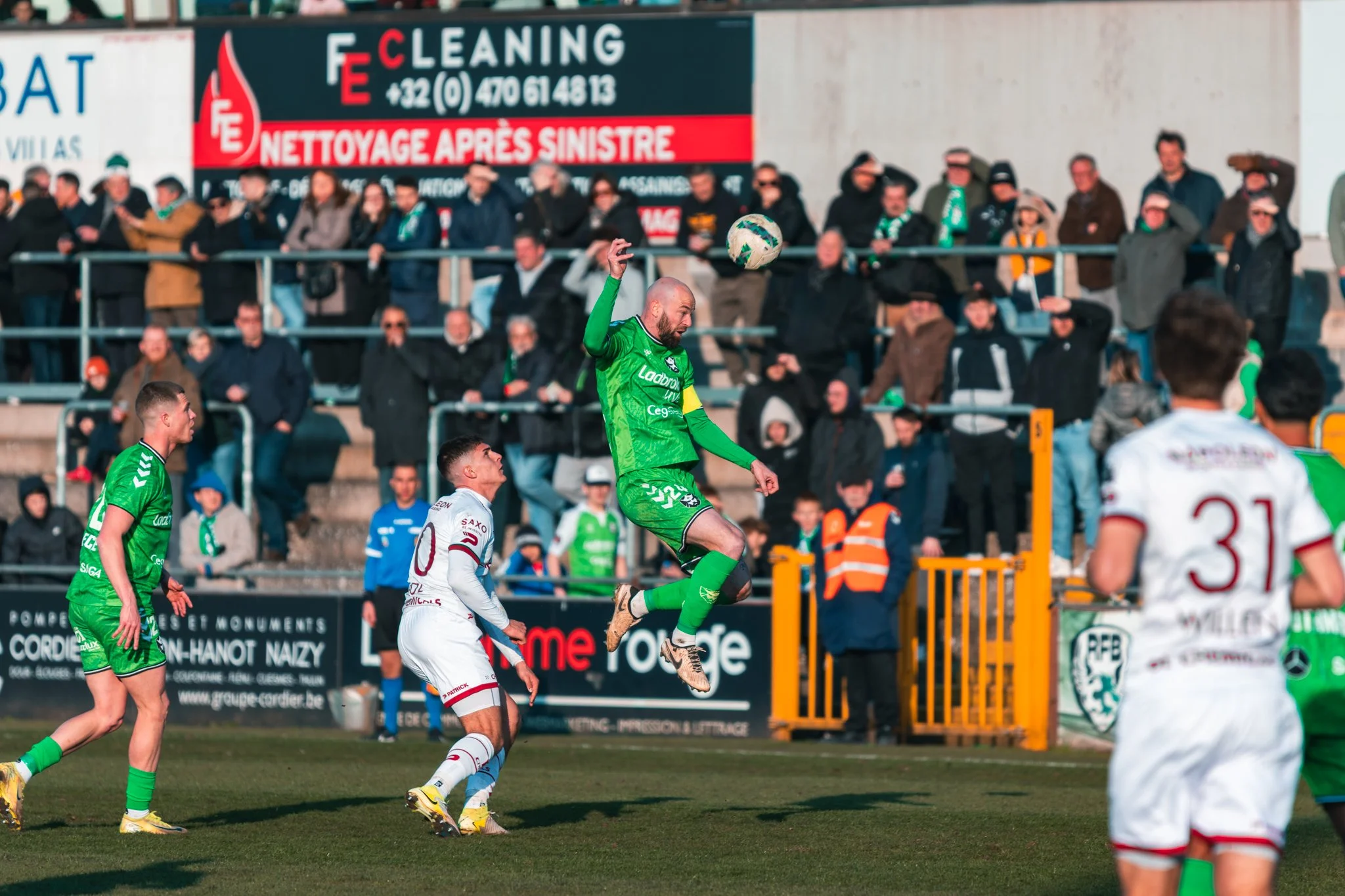 Un gardien de but en vert saute pour attraper un ballon lors d'un match de football. Des joueurs en blanc et vert sont visibles sur le terrain, avec des spectateurs derrière le grillage et une publicité en arrière-plan.