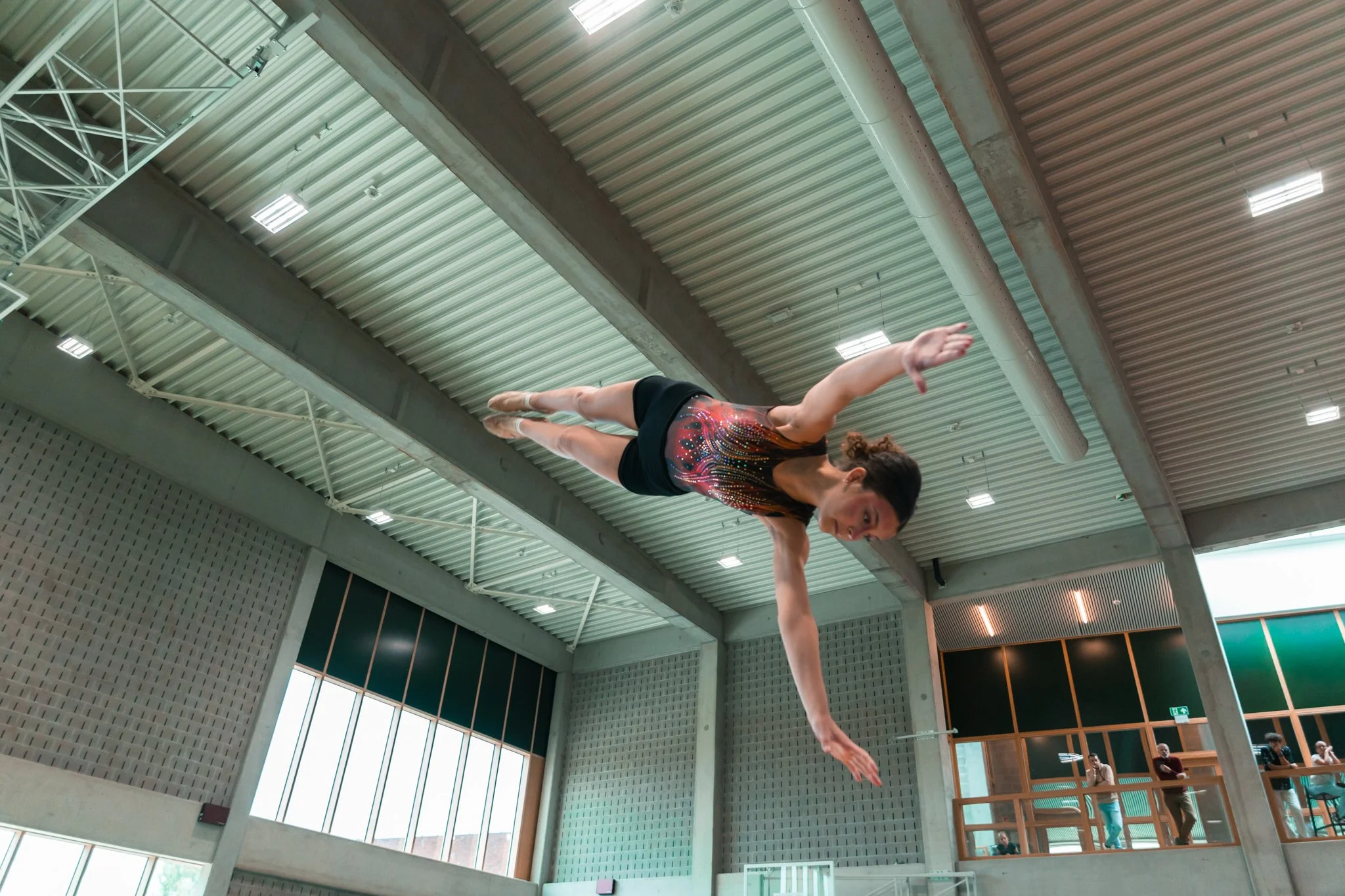 Une gymnaste effectue une acrobatie suspendue dans un espace intérieur, avec des spectateurs observant en arrière-plan.