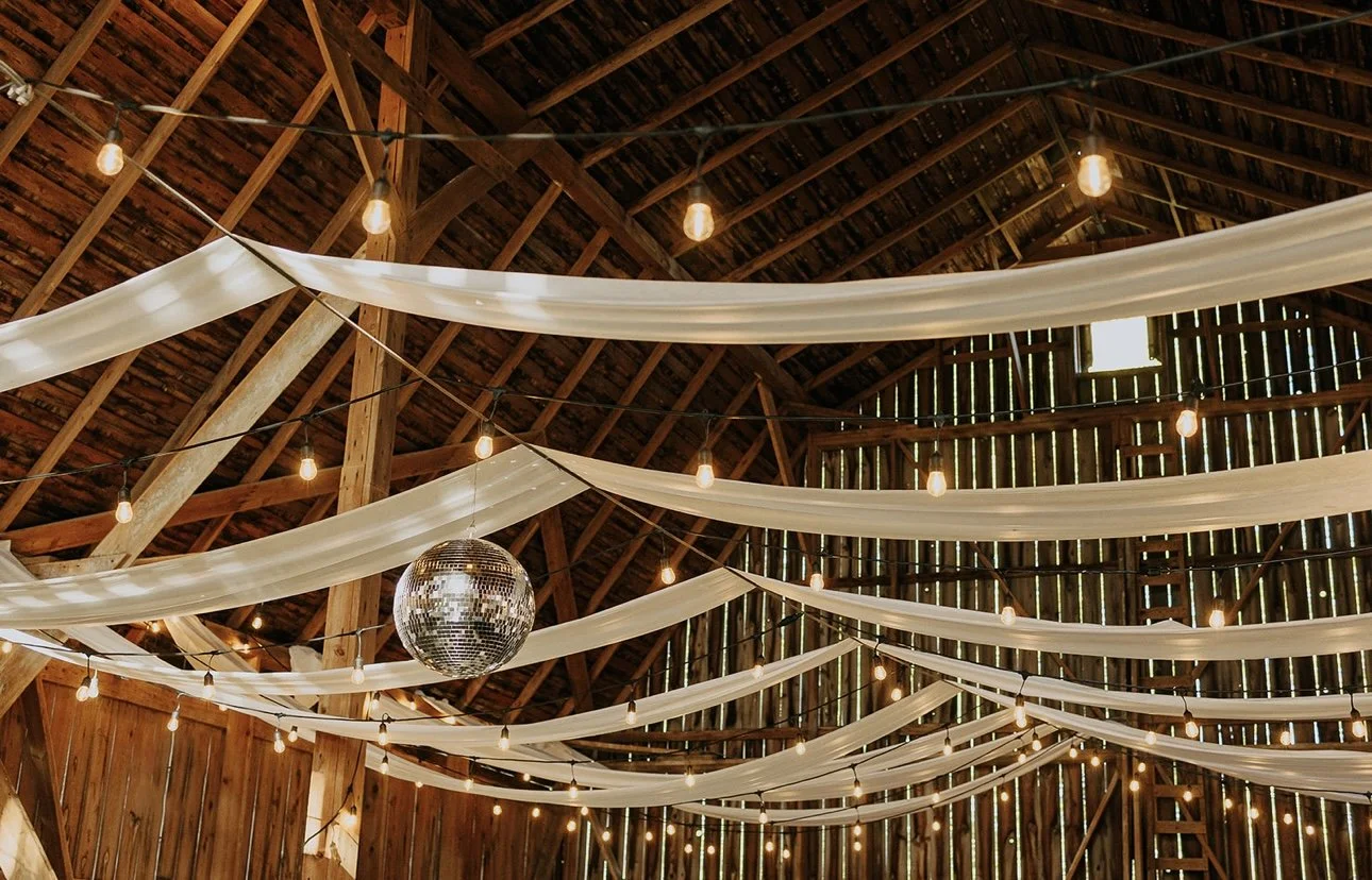 Decorative barn interior with string lights, white fabric drapes, and a disco ball hanging from the ceiling.
