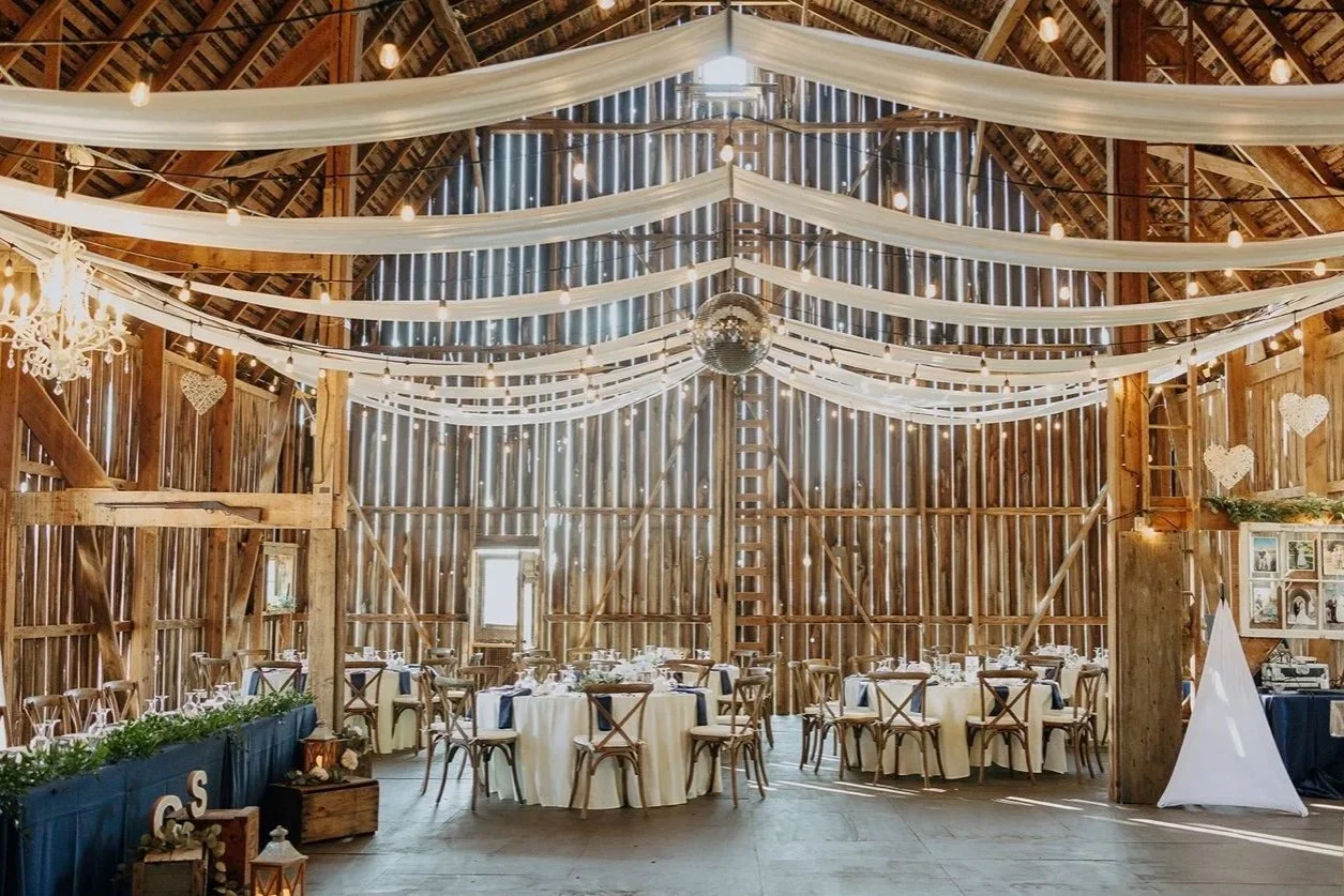 Interior of a rustic barn decorated for a wedding or celebration, with round tables, chairs, white tablecloths, string lights, fabric drapes, and a large disco ball hanging from the ceiling.