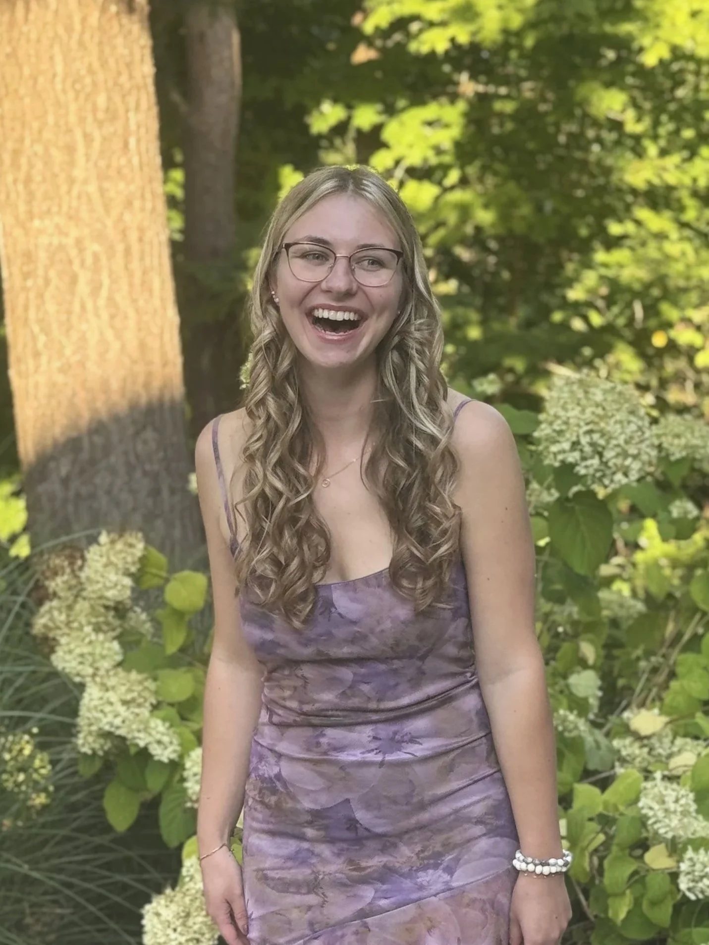 A young woman with long, curly blonde hair, glasses, and a big smile, wearing a purple leafy-patterned dress, standing outdoors in a green garden with trees and bushes.