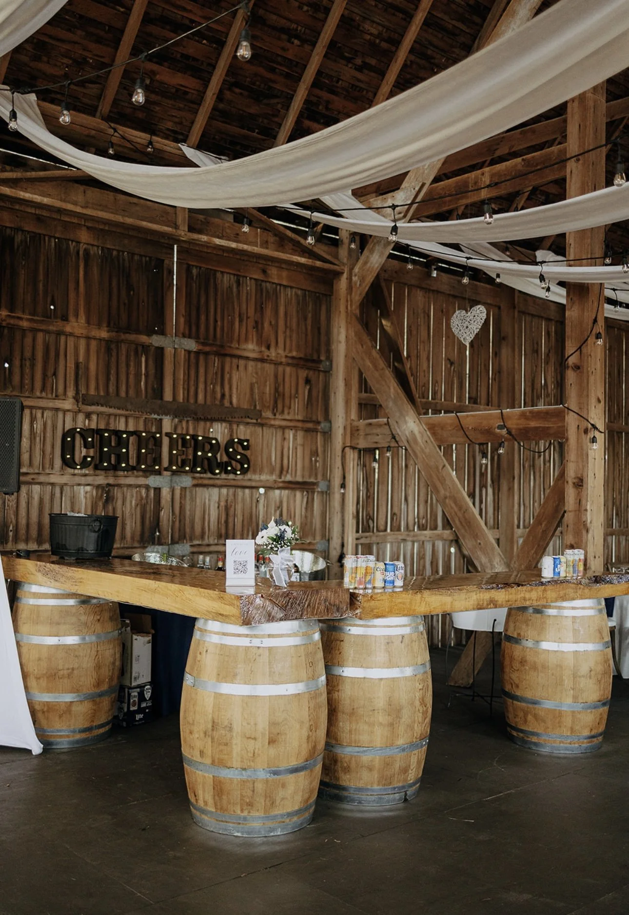 Wooden bar made from barrel staves with a thick live edge wooden countertop in a rustic barn setting, decorated with string lights and fabric drapes, with a sign spelling 'CHEERS' on the wall.