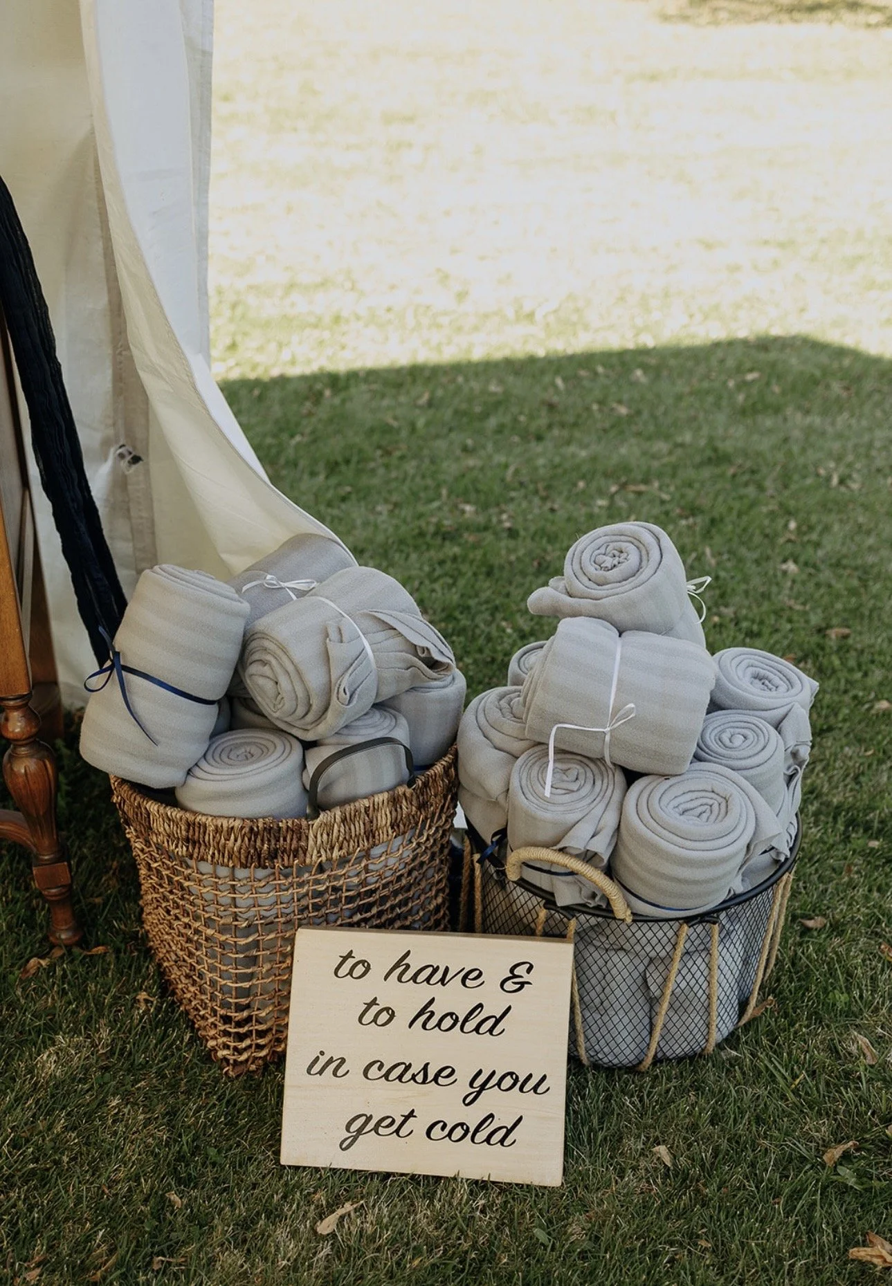 Multiple rolled-up white blankets in baskets with a sign that reads 'to have & to hold in case you get cold' outside on a grassy area near a tent or canopy.