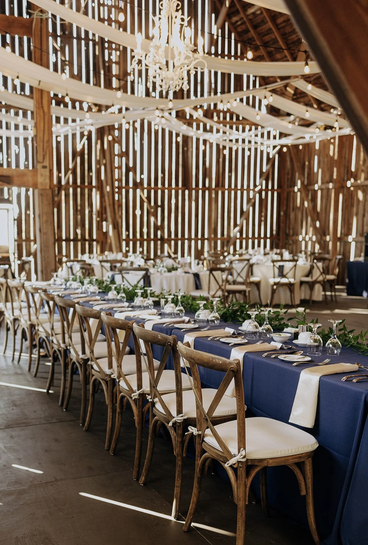 Elegant rustic wedding reception setup in a barn with long navy blue tablecloths, wooden cross-back chairs with white cushions, greenery garlands, and hanging wine glasses. A chandelier and string lights are suspended from the ceiling, with draped white fabric enhancing the rustic ambiance.