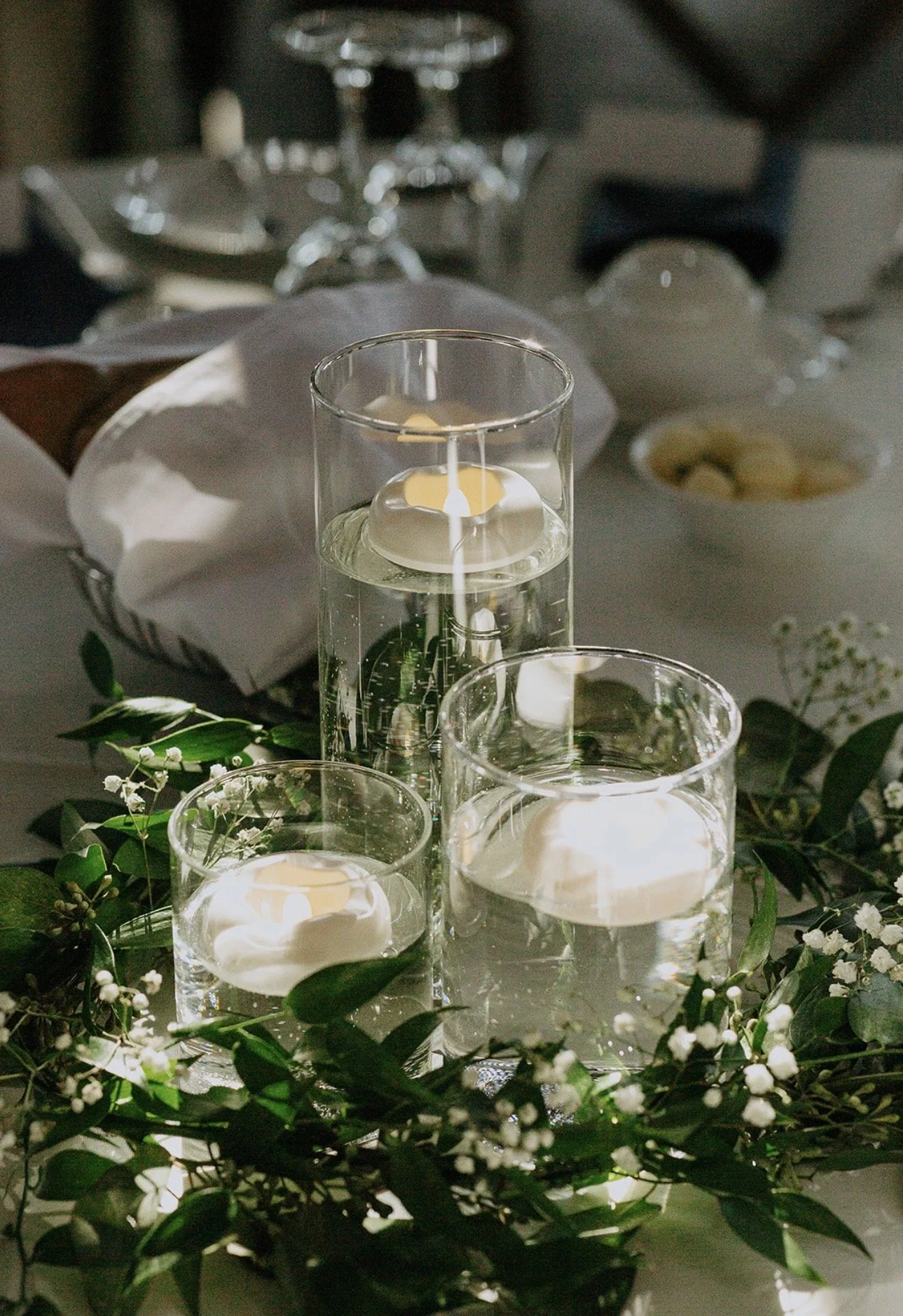 Three floating candles in glass holders float on water surrounded by greenery and small white flowers on a table, with a blurred background of additional tableware.