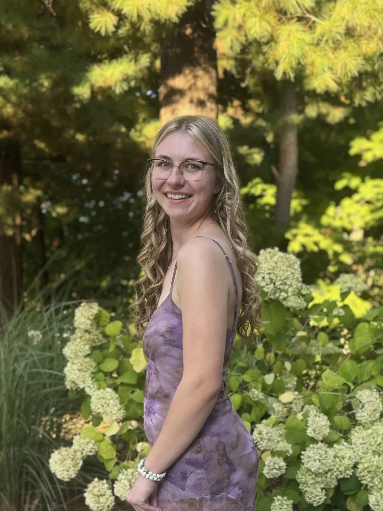 A young woman with blonde curly hair, glasses, and a purple floral dress, smiling and standing outdoors among green plants and white flowering bushes.