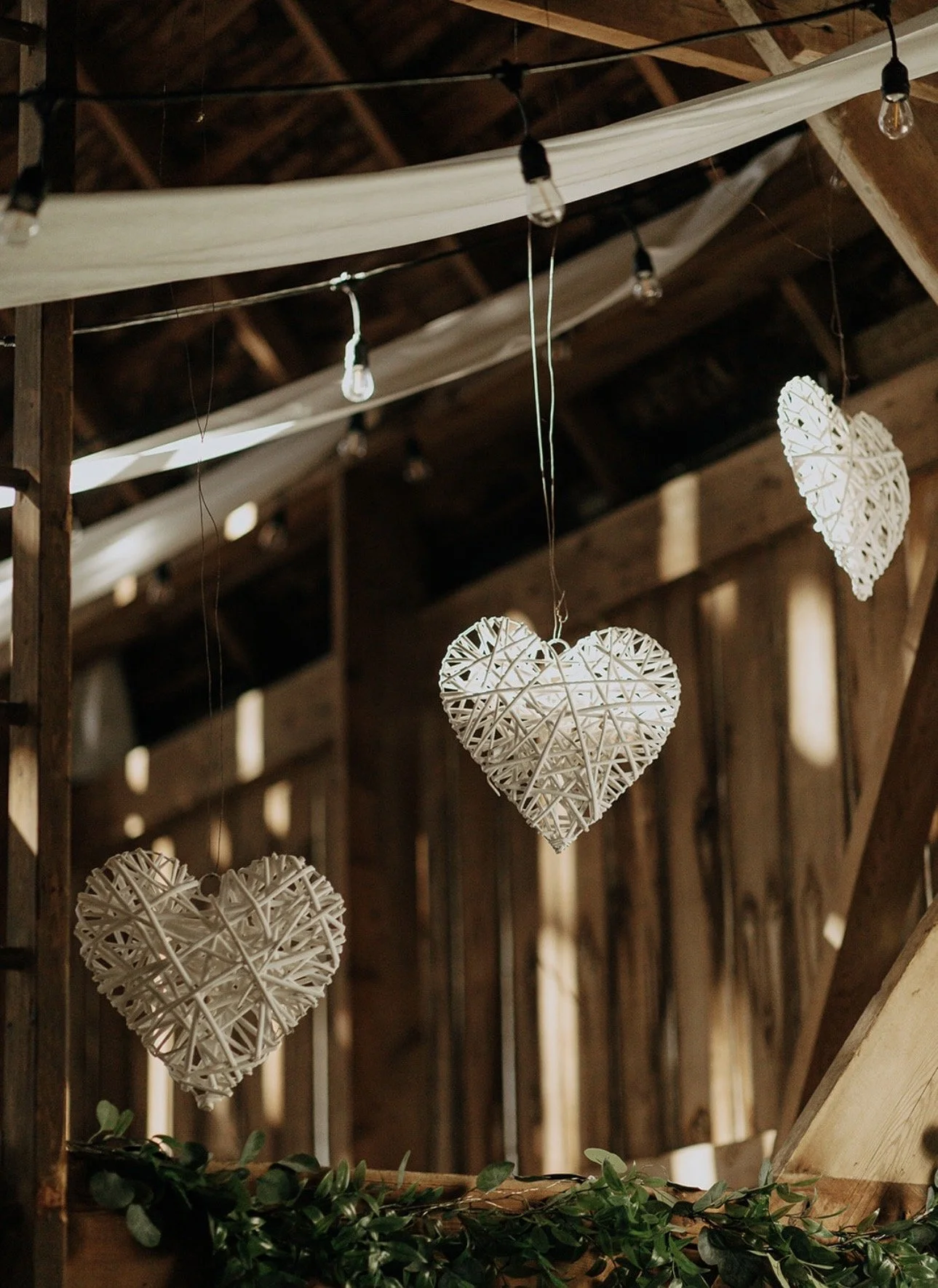 Hanging woven hearts and a heart-shaped decor in a rustic wooden barn with string lights and greenery.