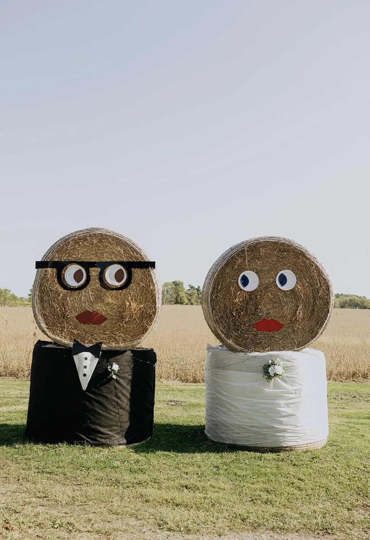 Two hay bales decorated as a bride and groom for a wedding. The hay bale on the left is dressed as a groom with glasses, a bow tie, and a tuxedo, while the hay bale on the right is dressed as a bride with a white wedding dress and a small bouquet. Both have paper eyes and red paper lips, standing on grass with a field and clear sky in the background.