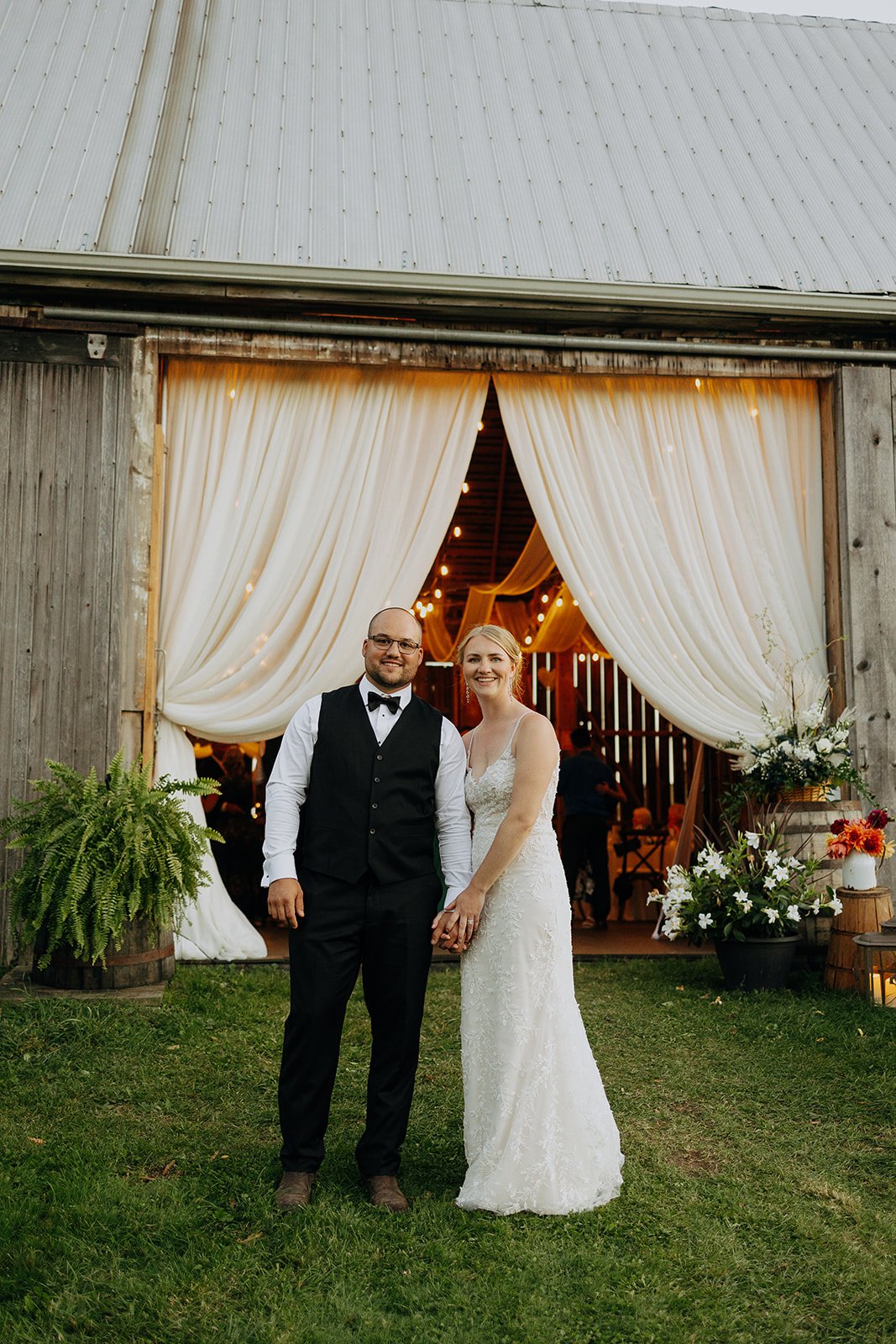 A newlywed couple is standing outside a rustic barn with white curtains and string lights, holding hands and smiling.