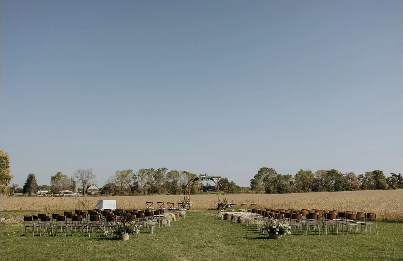 Outdoor wedding ceremony setup in a field with chairs arranged on either side of a grassy aisle, an arch at the front, and potted plants and flowers.