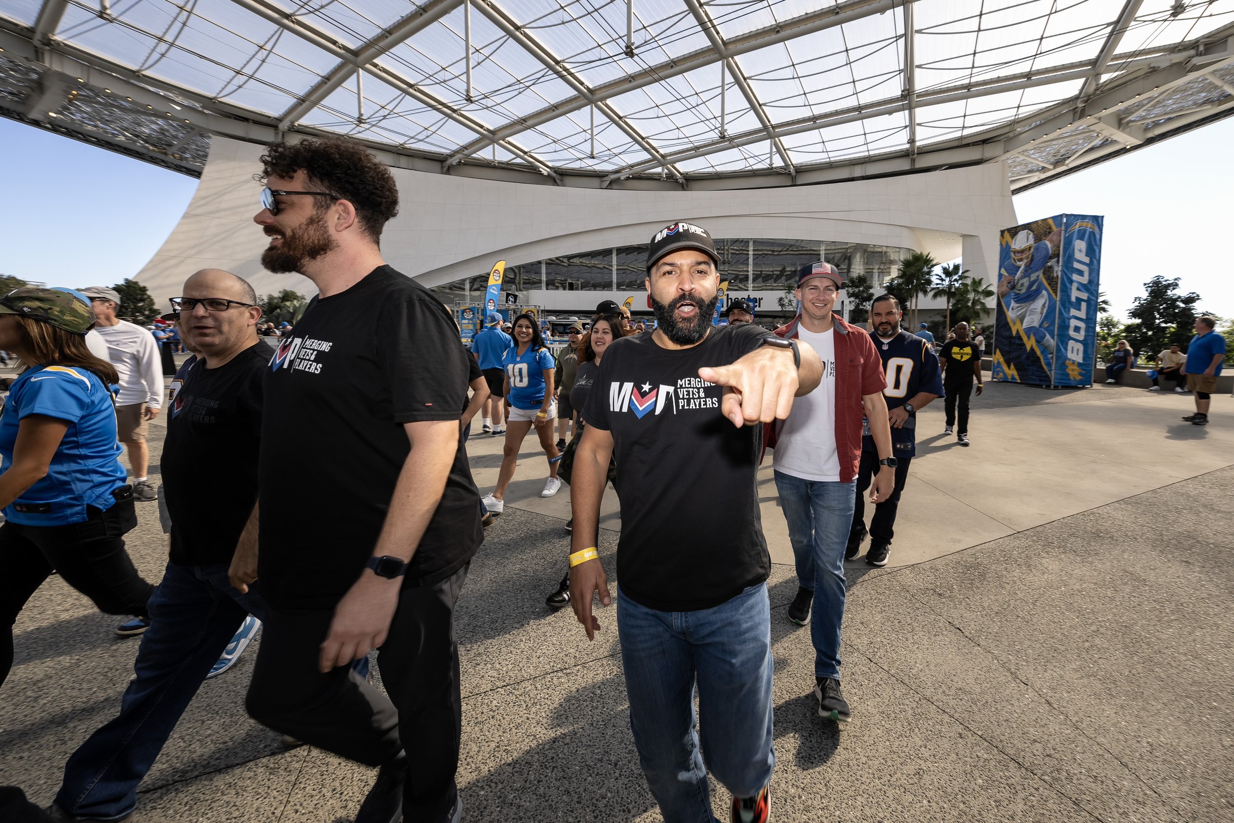 Group of people walking outside a sports arena, with a man in a black Scouts t-shirt pointing at the camera, others in team shirts and casual wear, a large graphic banner with a football player, and the arena with a glass roof in the background.