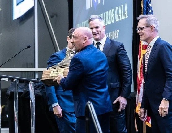 A group of men in business suits on stage at an award ceremony, with one man receiving a wooden plaque and all smiling.