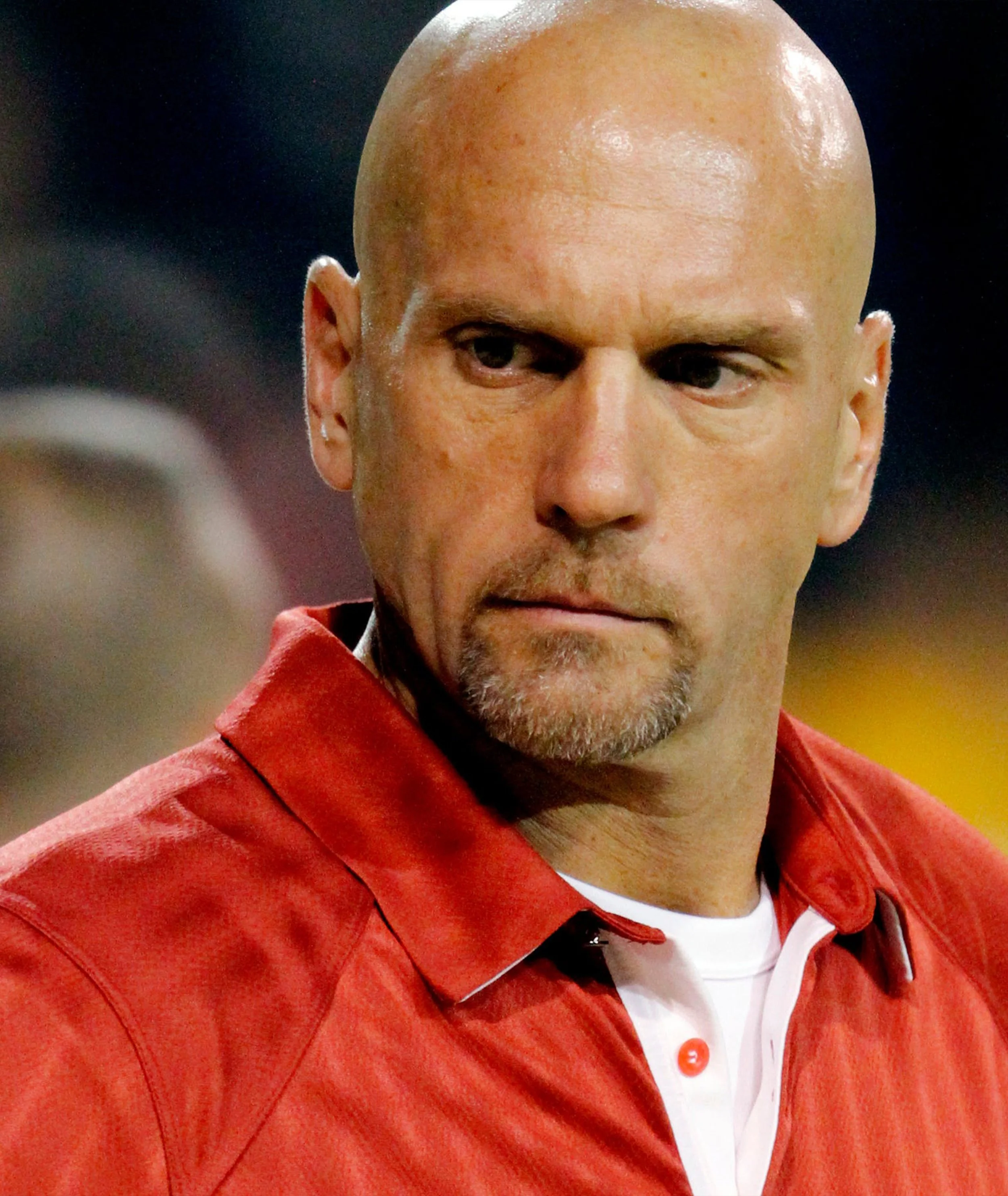 Bald man with goatee wearing a red collared shirt with white underneath, looking serious.
