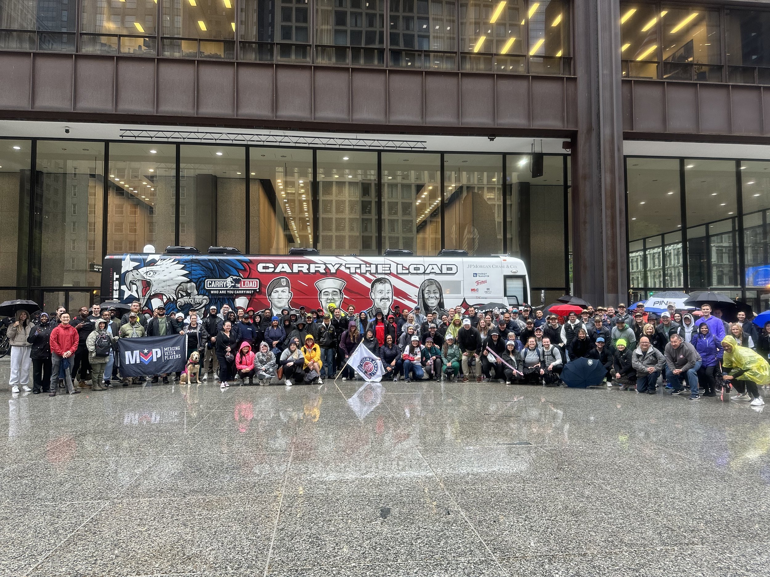 A large group of people standing in front of a city building on a rainy day, with umbrellas and raincoats. They are posing for a photo in front of a bus that has patriotic and sports-related graphics. Some people are holding flags and banners.