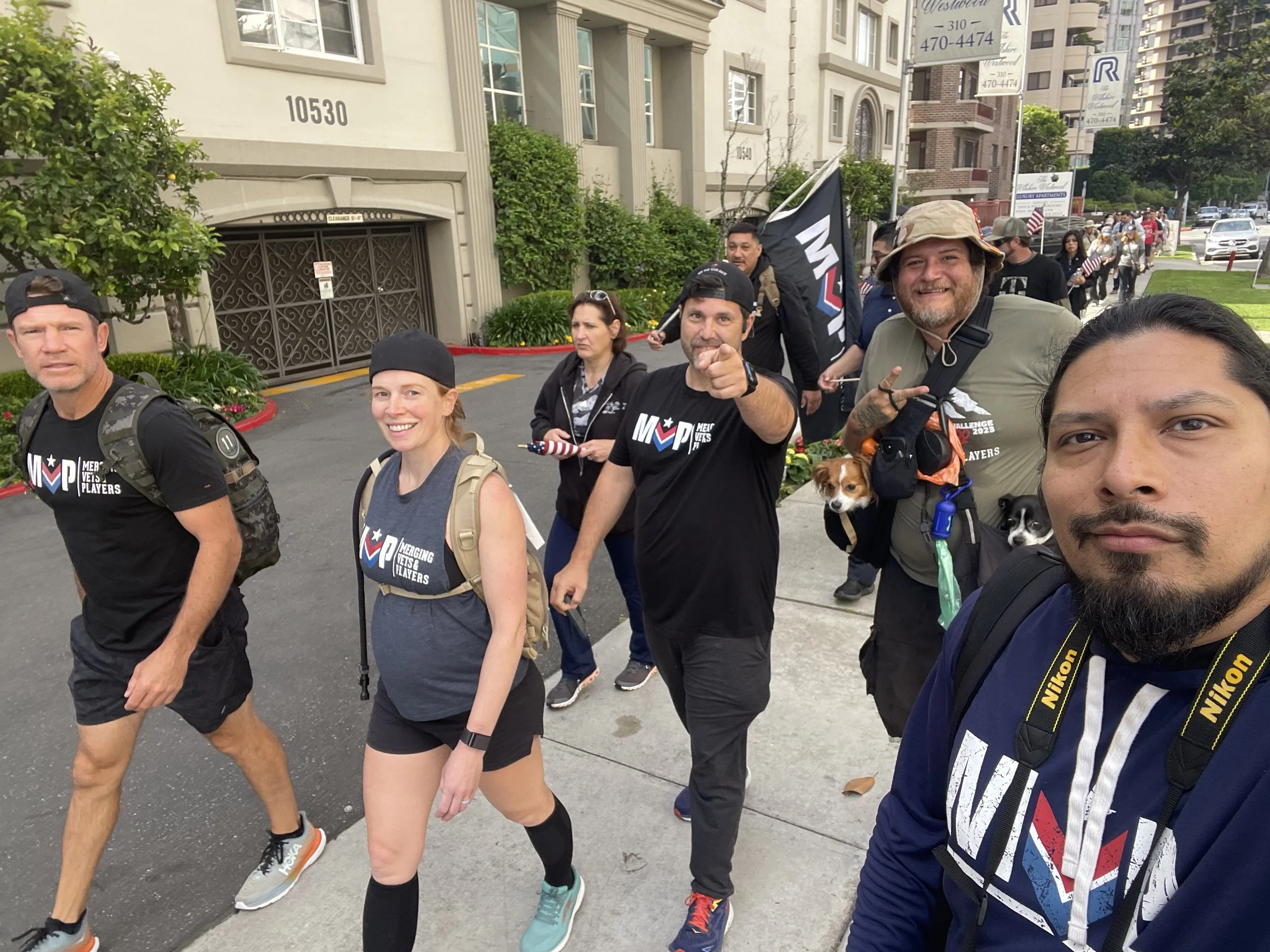 Group of people participating in the Murph Veterans challenge walk, wearing clothing with event logos, some carrying backpacks and dogs, walking along a sidewalk in a residential neighborhood.