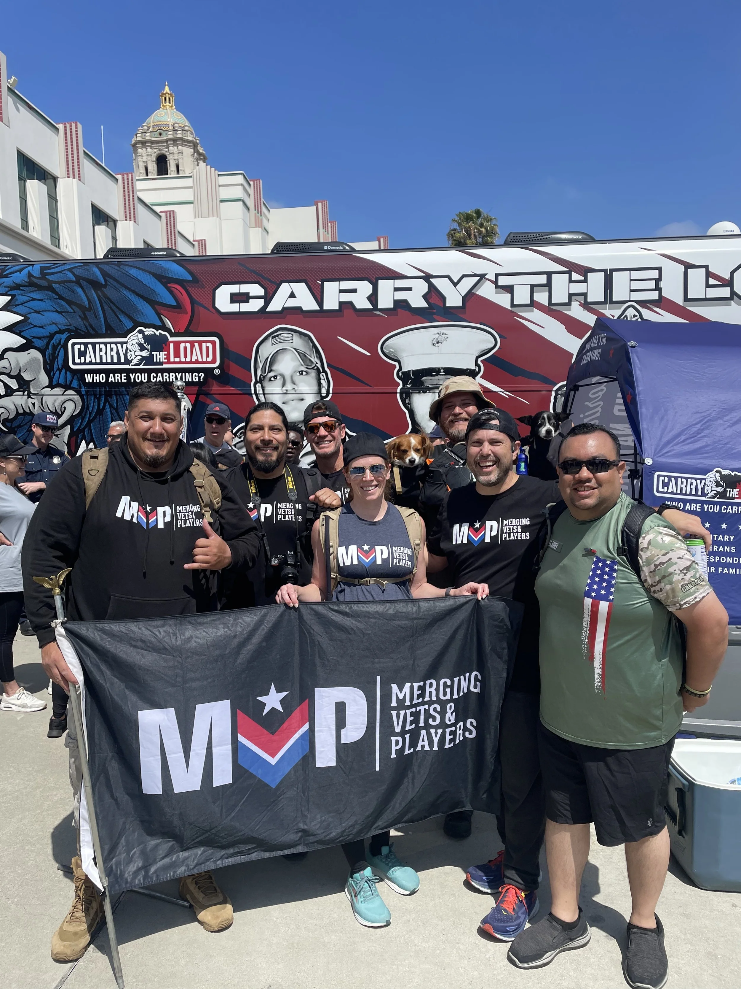 Group of people holding a banner that reads 'Merging Vets & Players', standing in front of a large vehicle with 'Carry the Load' branding, some wearing sunglasses, military and civilian clothing, and a few with dogs, on a sunny day outside a historic building.