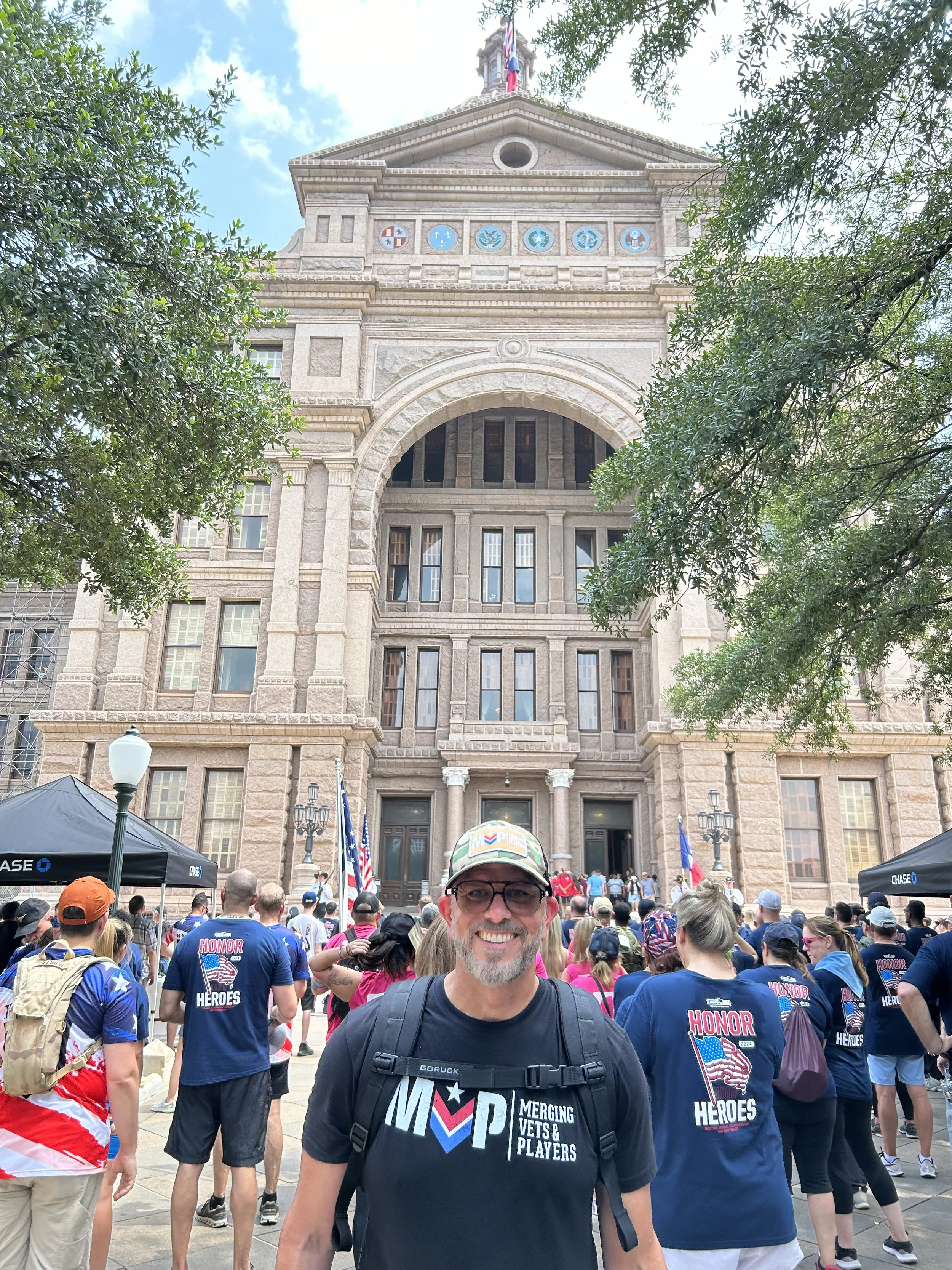 Man smiling at a public gathering in front of a historic building with flags, surrounded by people wearing patriotic shirts and hats.