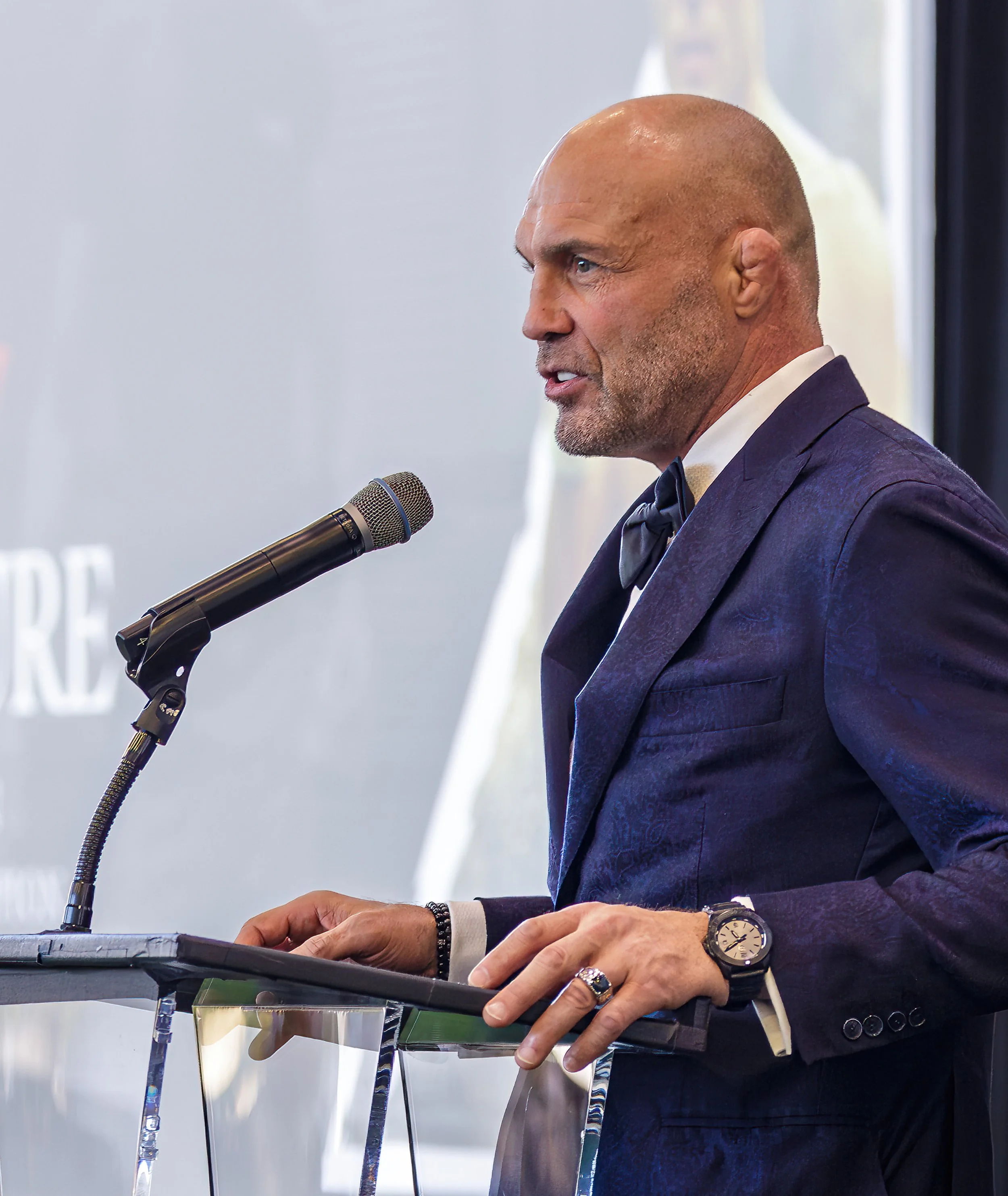 A man in a dark blue tuxedo speaking at a podium with a microphone, wearing a watch and rings.