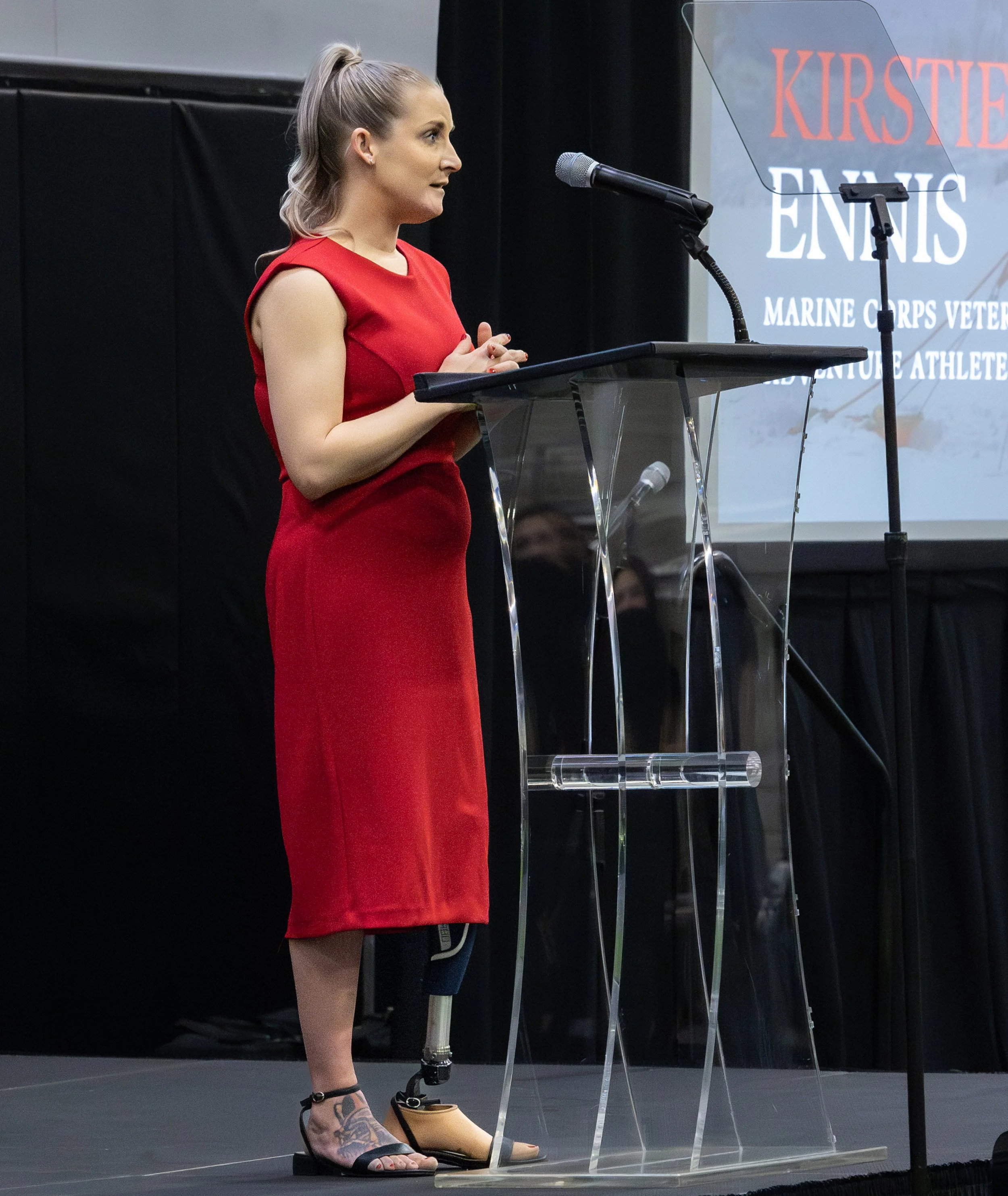 Woman in a red dress standing at a podium presenting at an event with a projection screen behind her displaying her name and title.