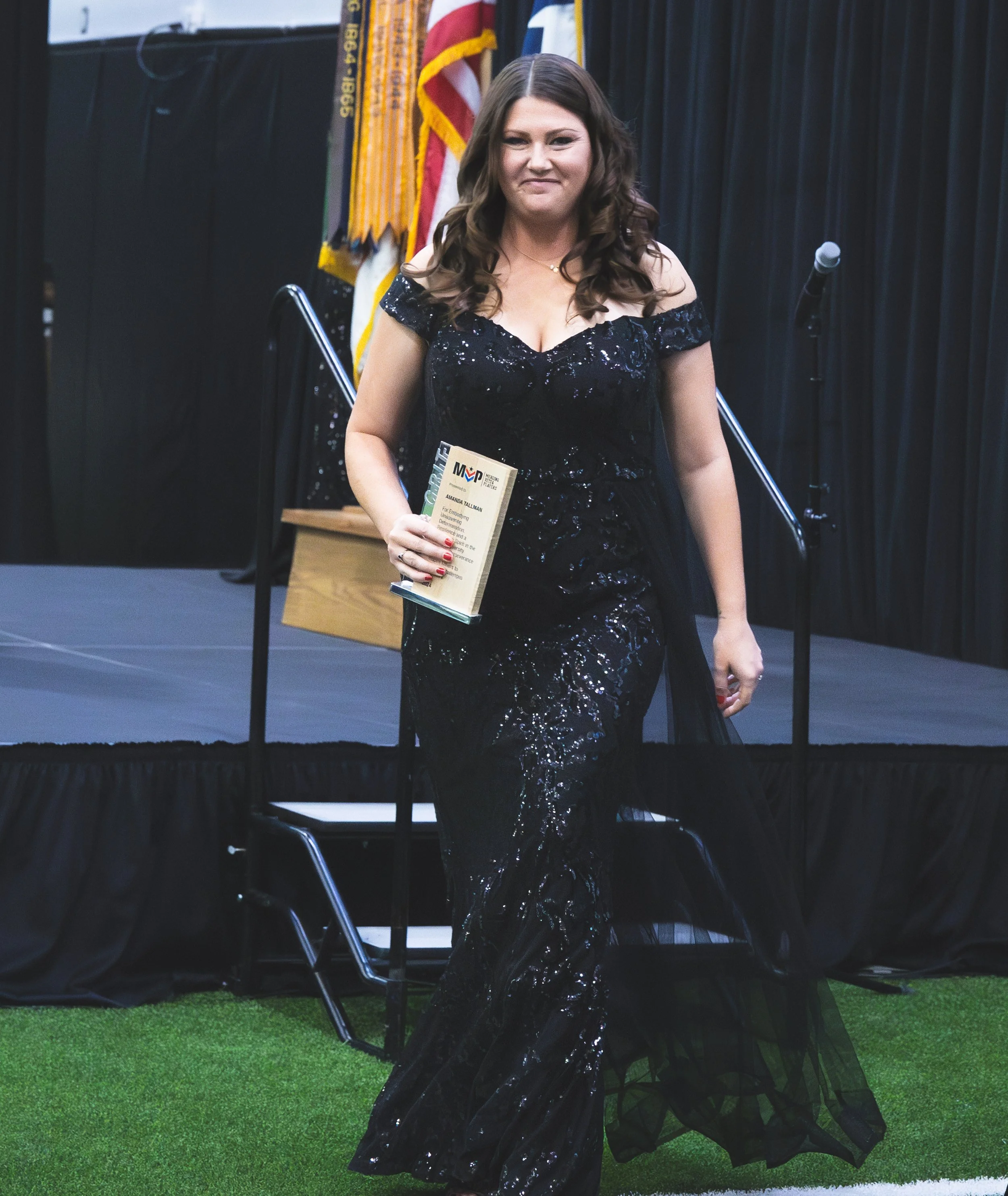 Woman in a black sequined dress holding a plaque or award, standing on a stage with black curtains and flags in the background.