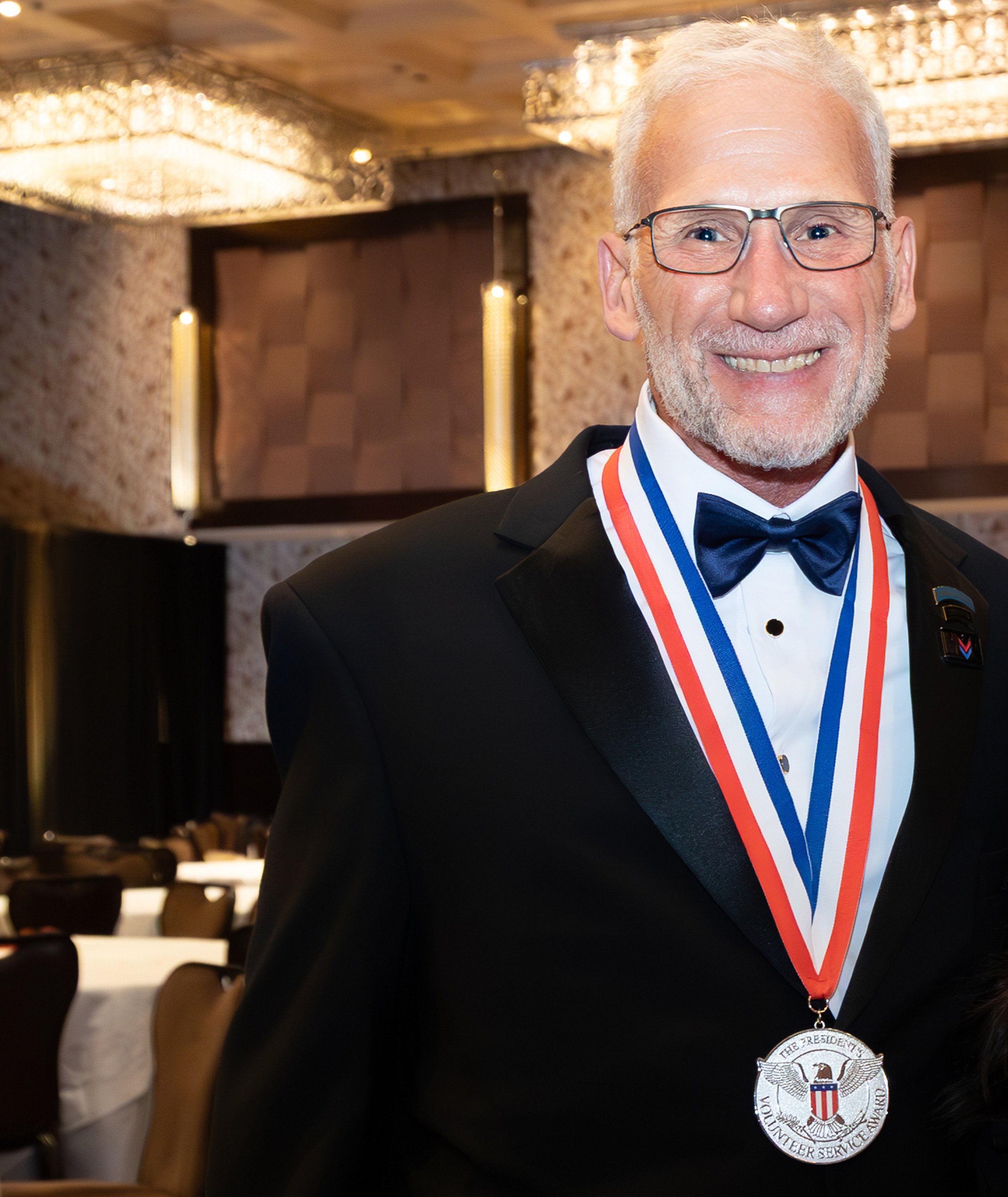 A smiling elderly man in a tuxedo with a bow tie, wearing a medal on a red, white, and blue ribbon, at a formal event.