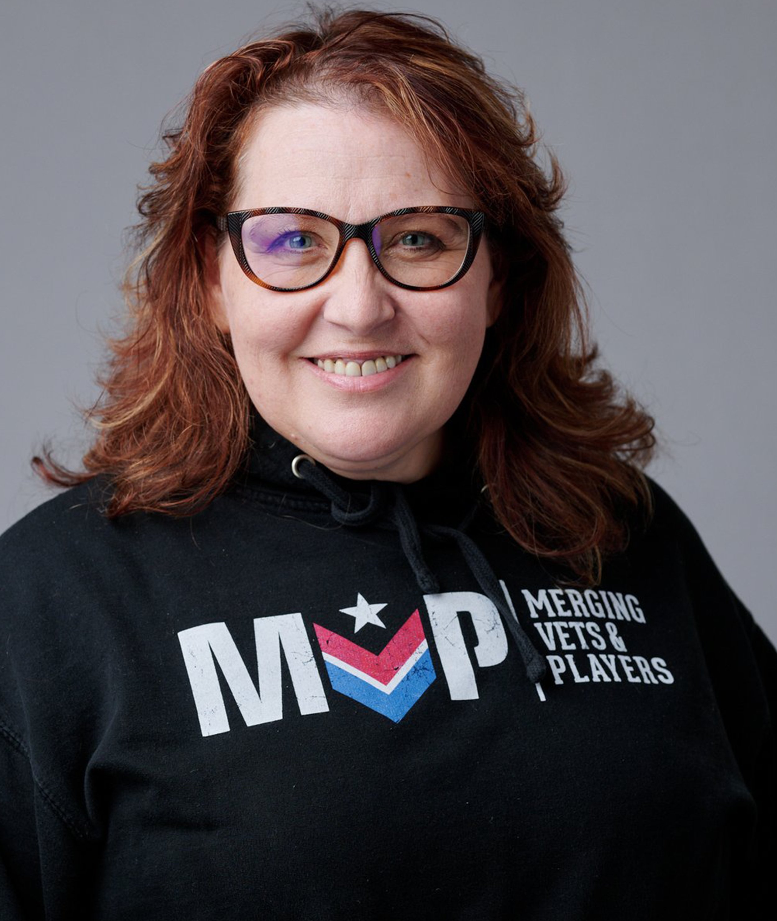 A woman with red hair, glasses, and a black hoodie smiling at the camera. The hoodie has text that reads 'MVP Merging Vets & Players' with a star and chevron logo.