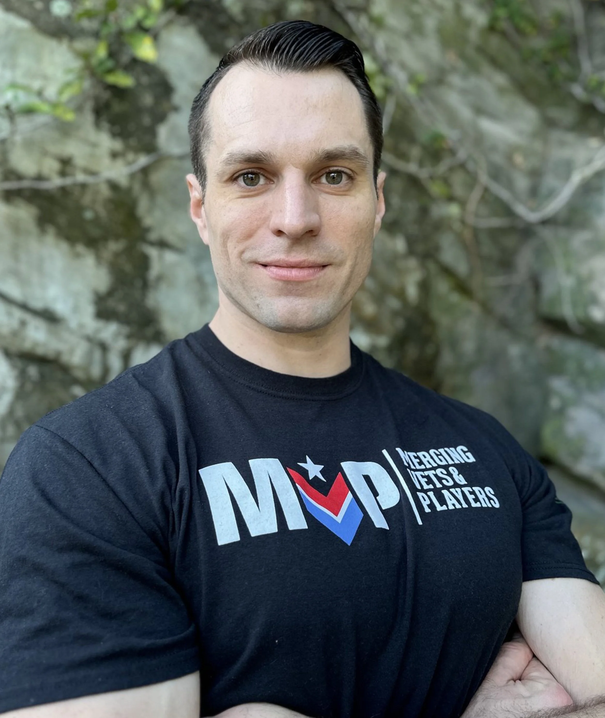 A man with dark hair and light skin standing outdoors in front of a rocky background, wearing a black T-shirt with a logo and text.