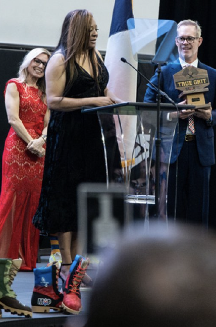 A woman in a black dress speaking at a podium while a man in a suit and glasses holds an award and a plaque that reads 'True Grit.' A woman in a red dress stands behind them, smiling.