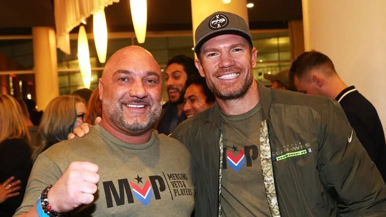 Two smiling men at an indoor event, one with a bald head and beard wearing a gray T-shirt, the other with a beard and wearing a cap and jacket, surrounded by other people.