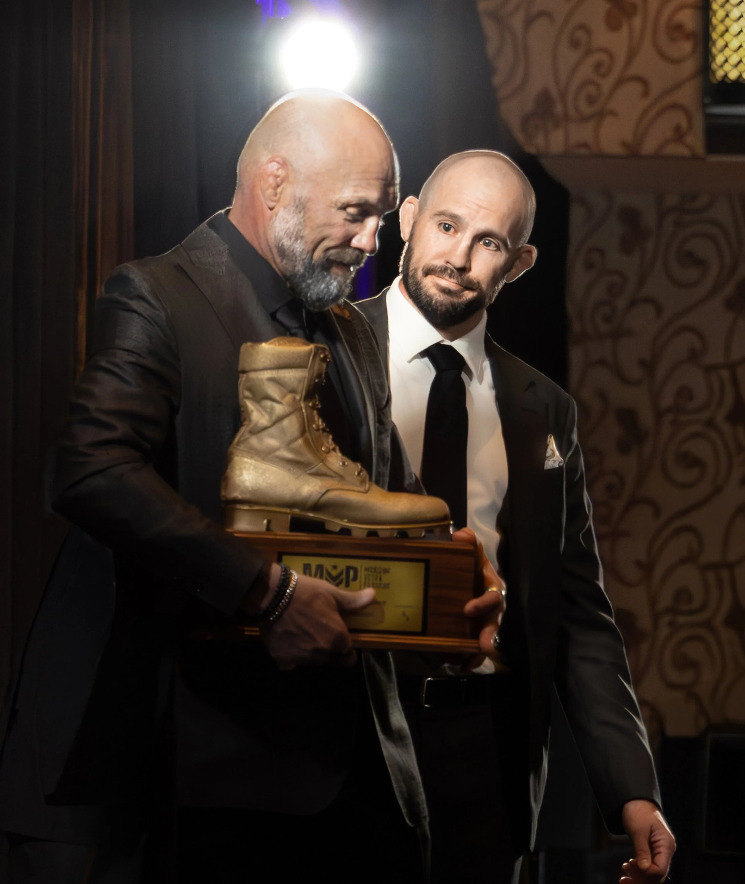 Two men in formal suits, one with a beard, are standing at an awards ceremony. The man on the left is holding a trophy shaped like a gold work boot mounted on a wooden base.