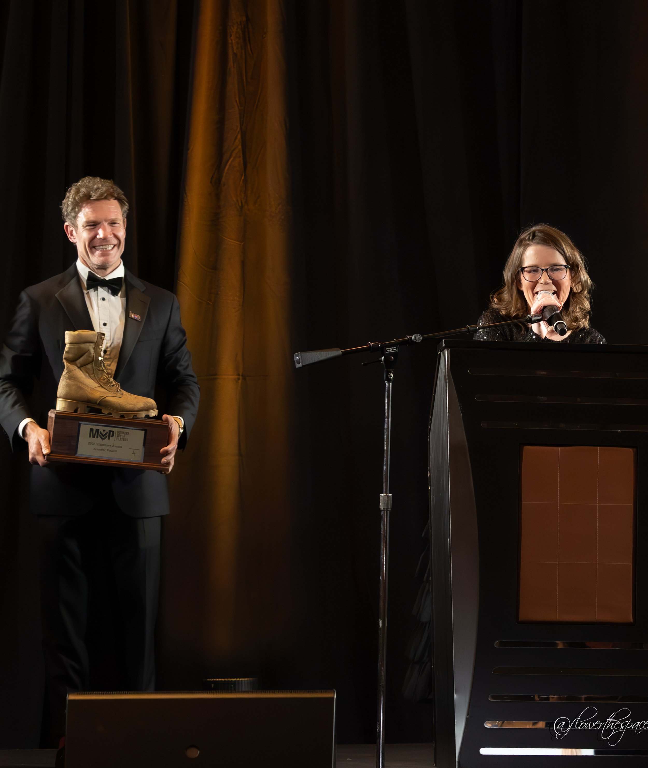 A man in a tuxedo holding a wooden award plaque with a tan hiking boot on top, and a woman singing into a microphone at a podium during an award ceremony on a stage with dark curtains.