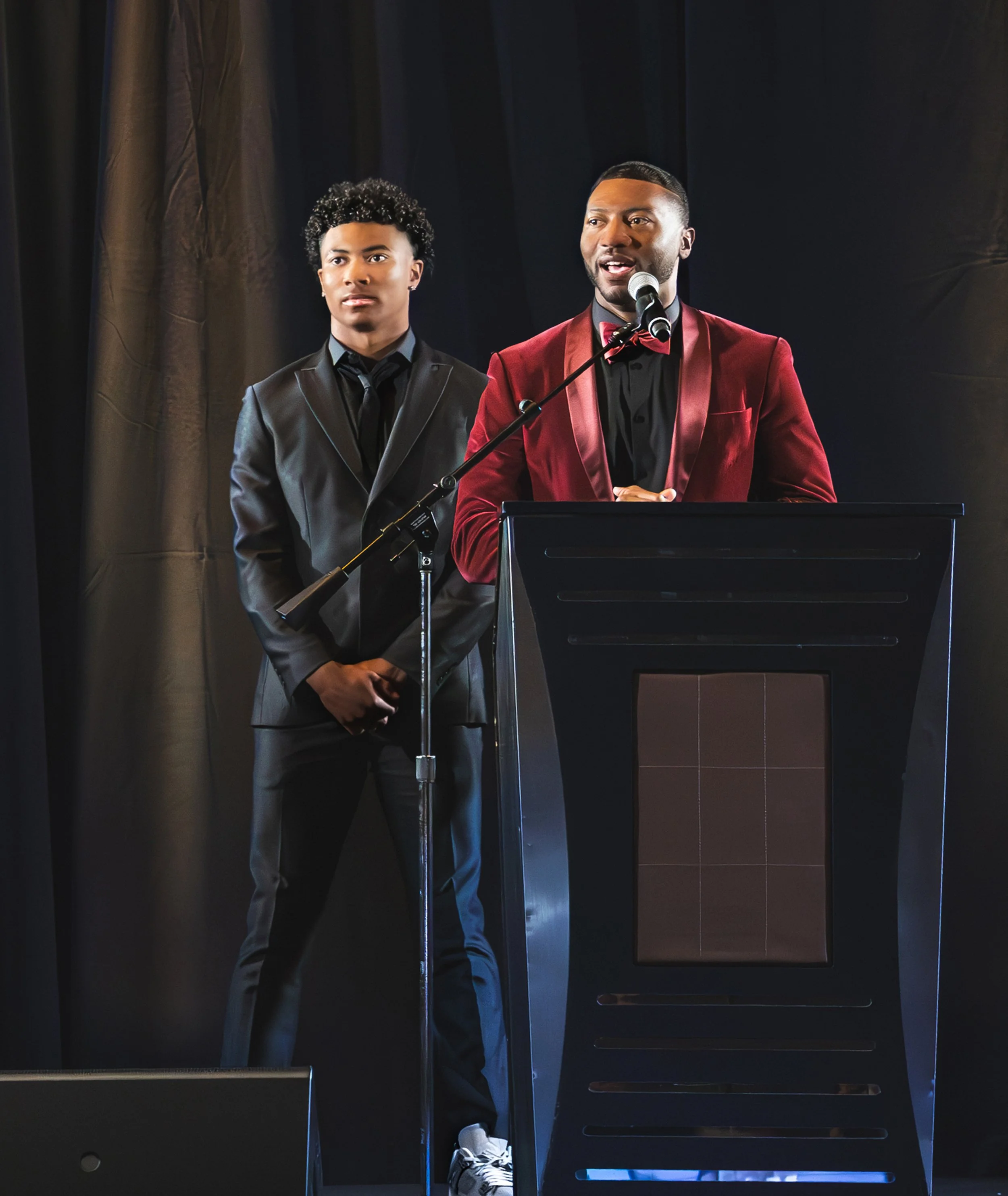Two men in suits speaking at a podium during a formal event, with a dark curtain in the background.
