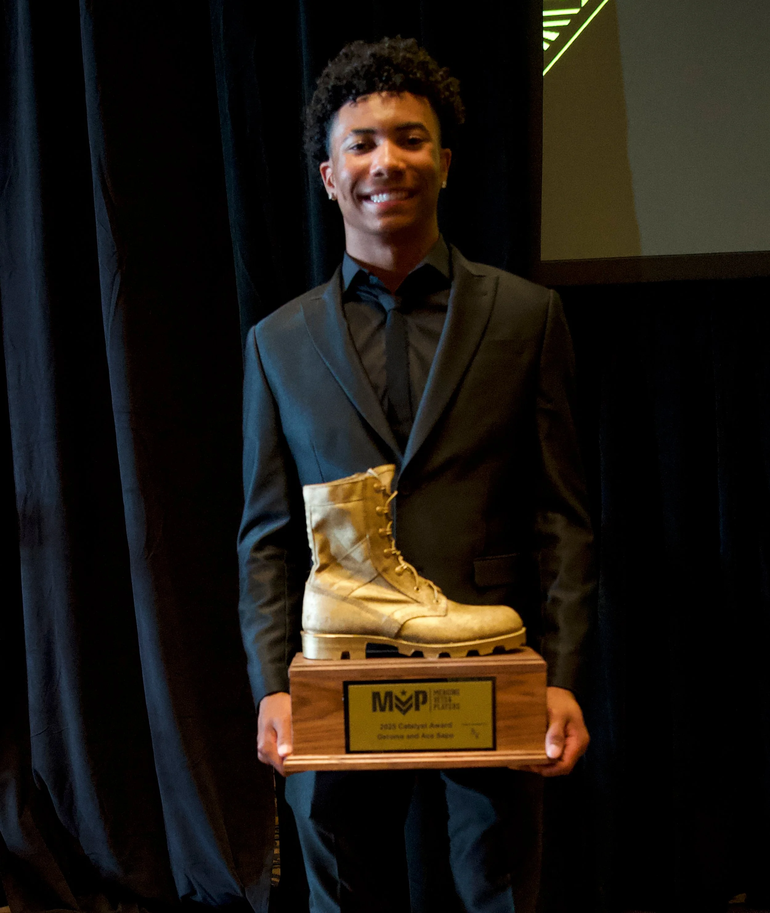 A young man in a black suit holding a wooden award plaque with a gold boot on it at an indoor event.