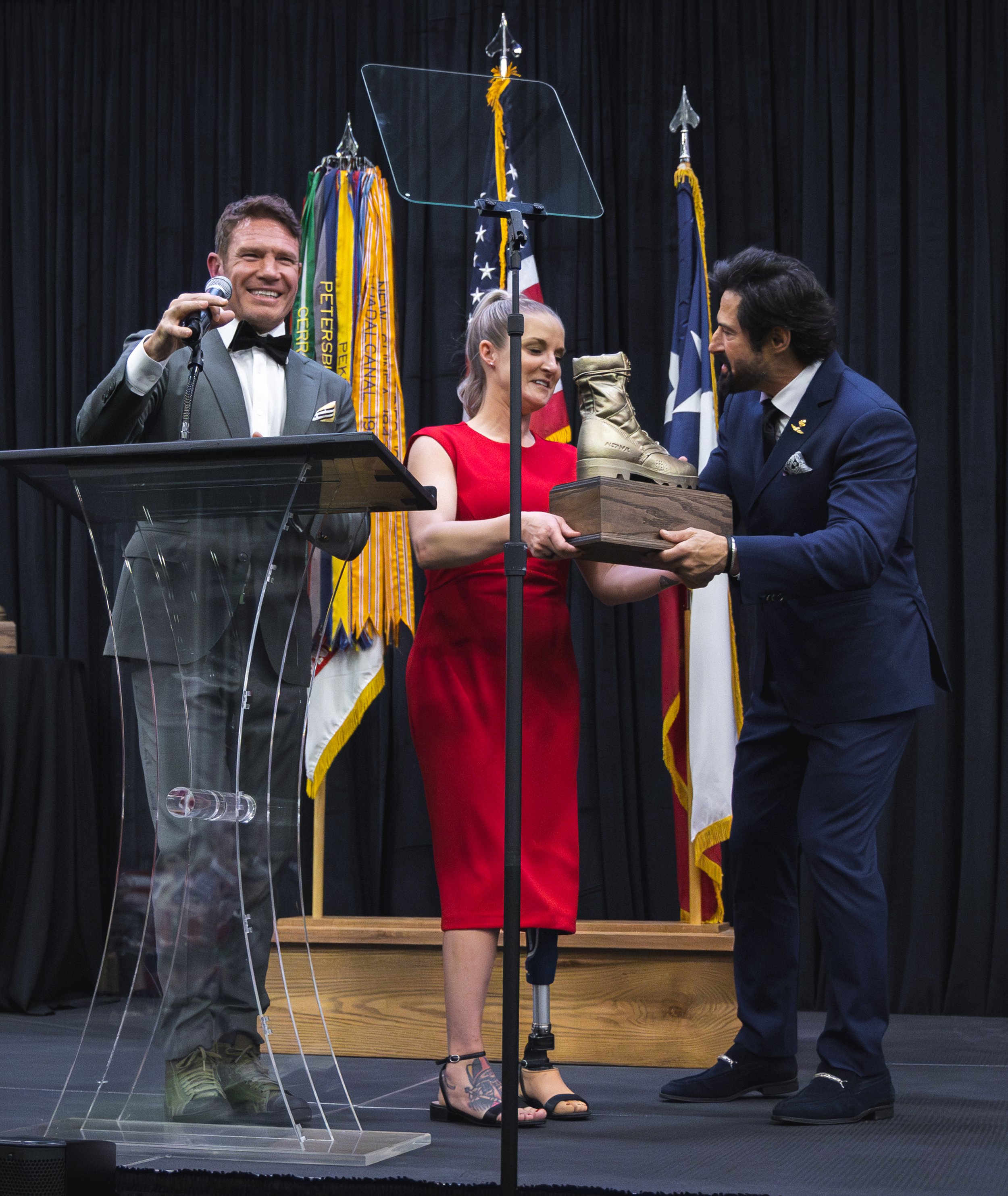 Woman in red dress receiving a pair of golden boots on stage during an awards ceremony, with man in grey suit and another man in dark blue suit presenting the boots. Flags are in the background.