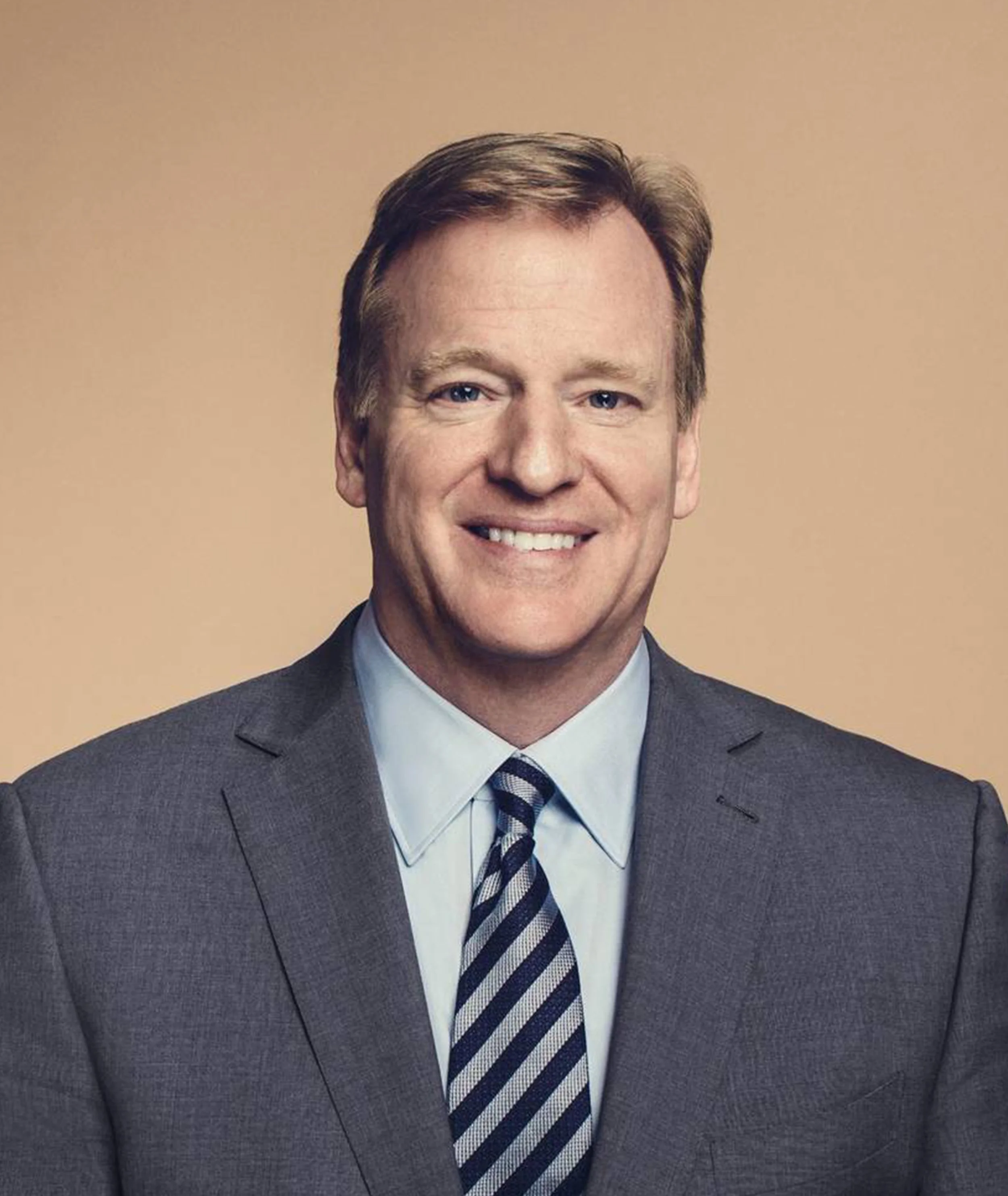 A man in a gray suit and striped tie smiling with a beige background.