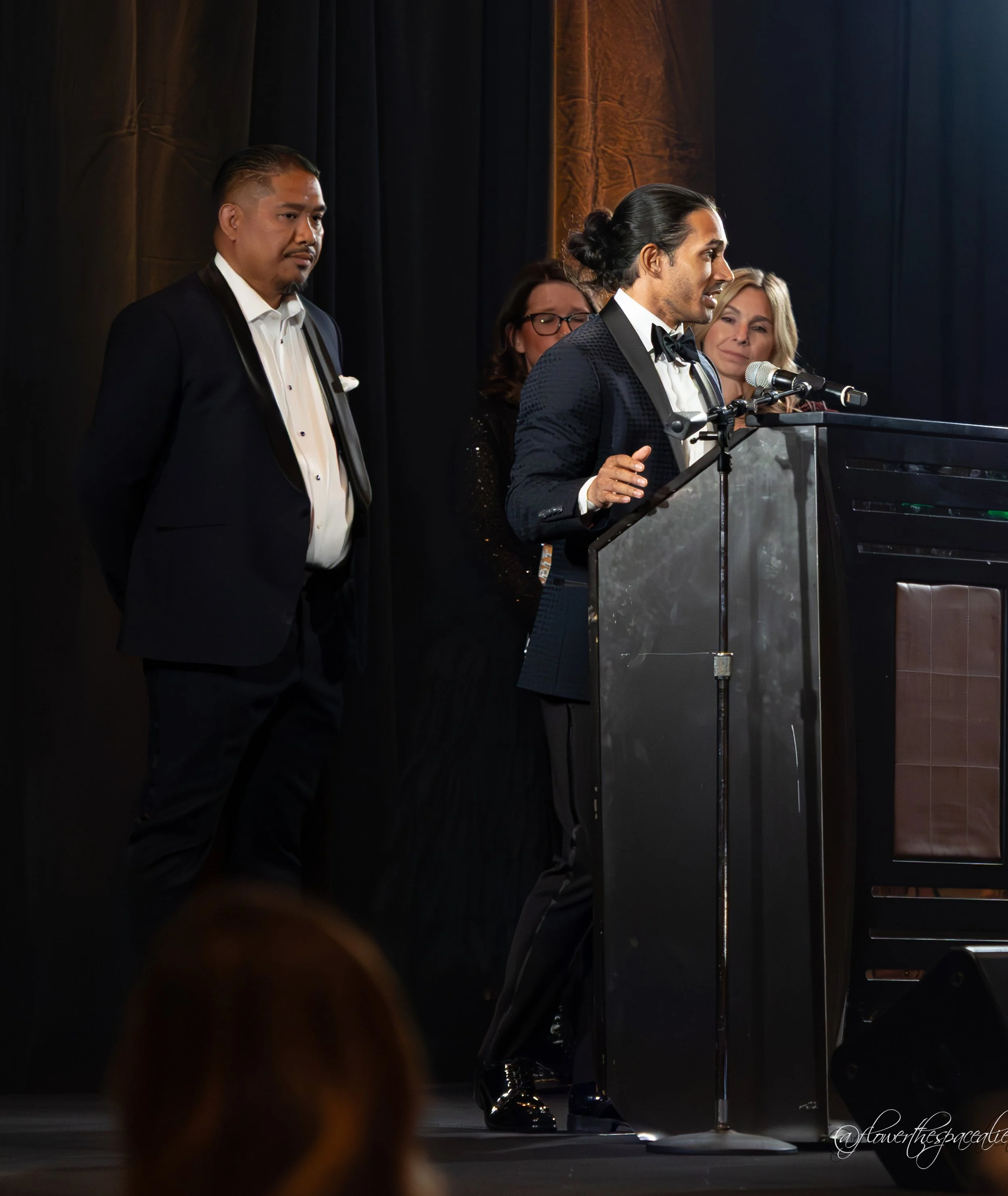 Man with long dark hair tied back and wearing a tuxedo speaking at a podium with a microphone, accompanied by several women and a man in tuxedos standing behind him on stage.