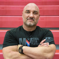 A bald man with a beard wearing a black t-shirt and a watch, standing in front of red bleachers.