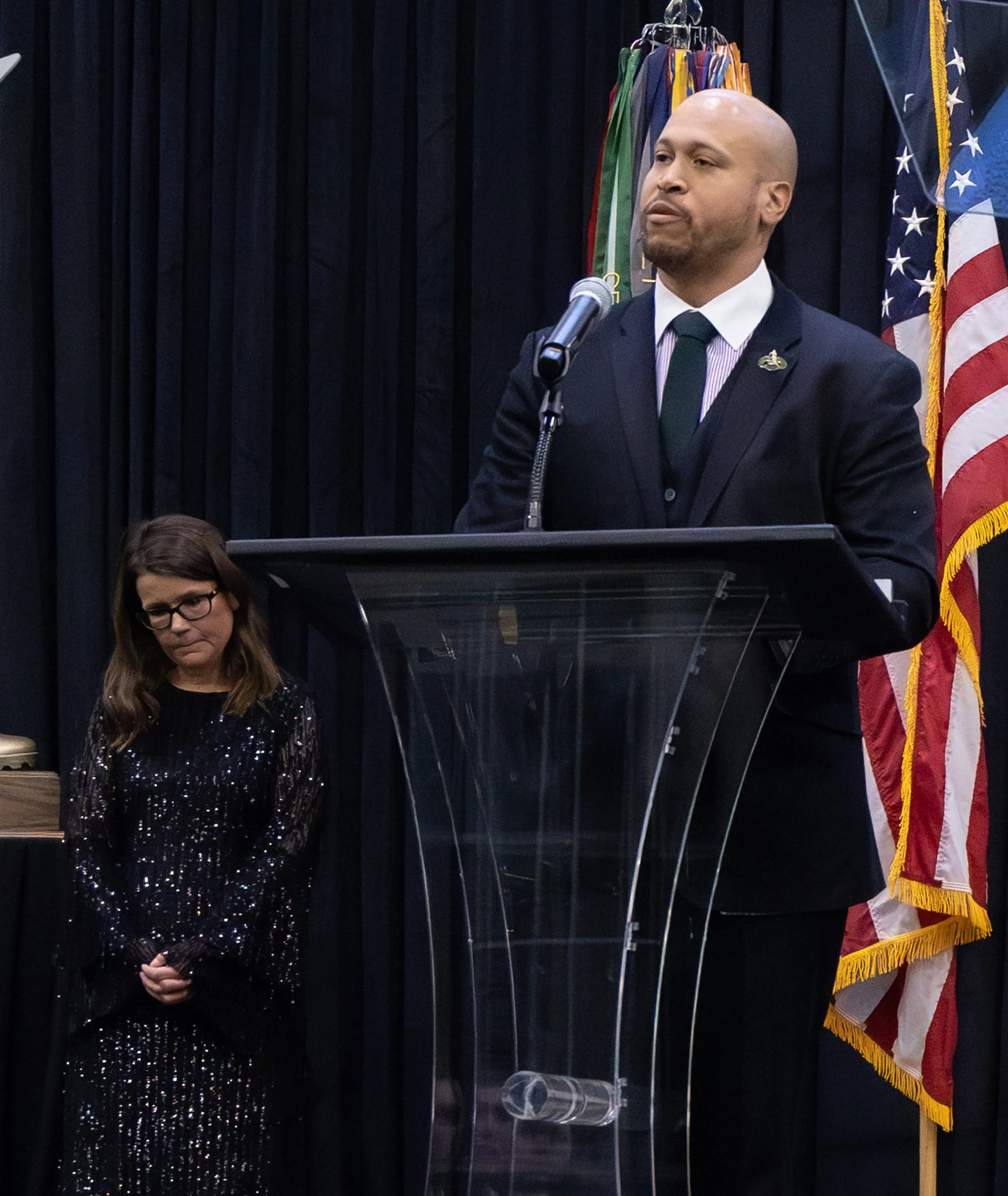 A man in a black suit and tie standing at a lectern, speaking into a microphone, with a woman in a black sequined dress standing with his head bowed, behind a backdrop of dark curtains, flags, and a coat of arms.